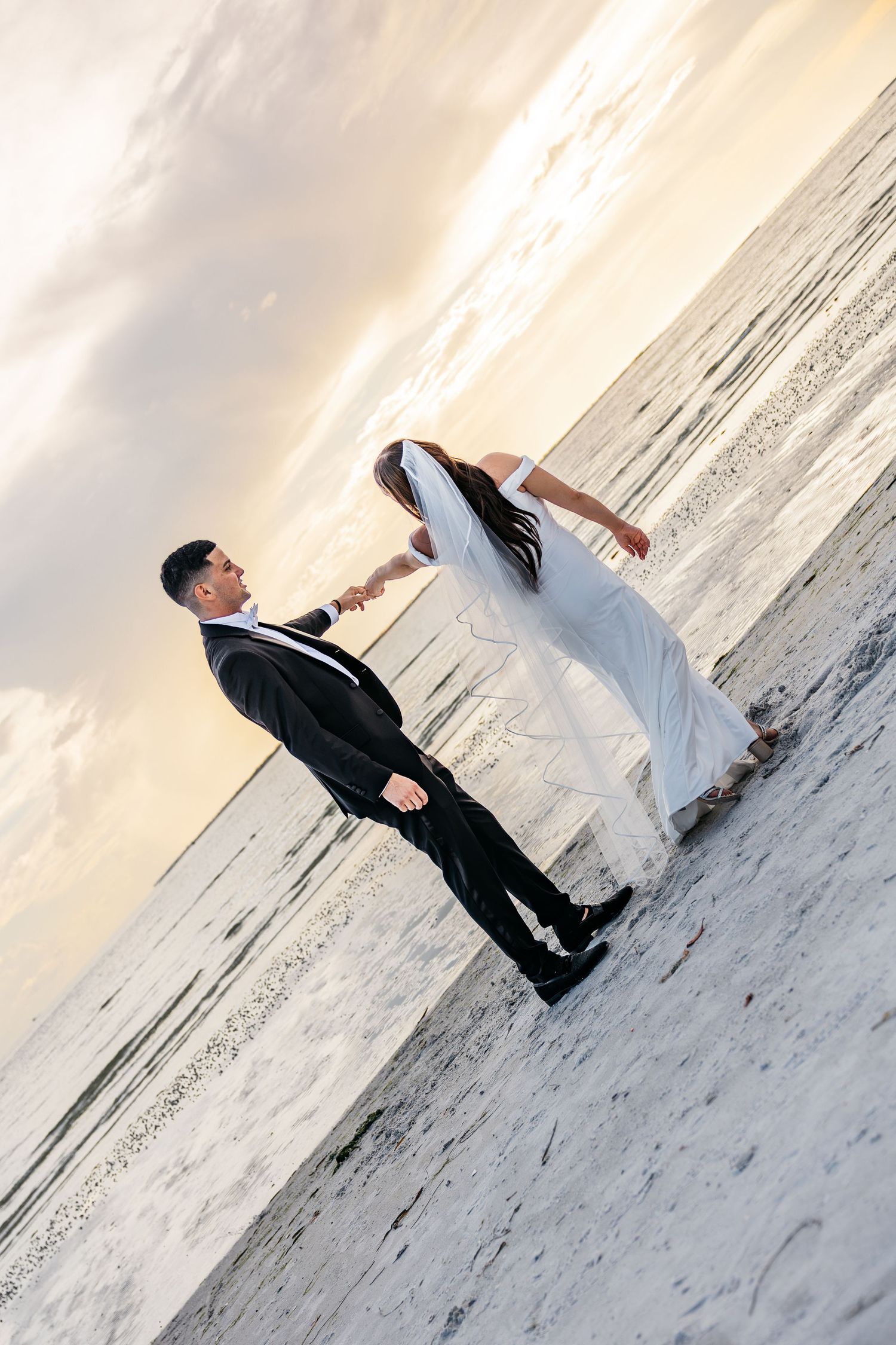 Bride and groom holding hands on a beach at sunset, wearing wedding attire with ocean waves in the background.