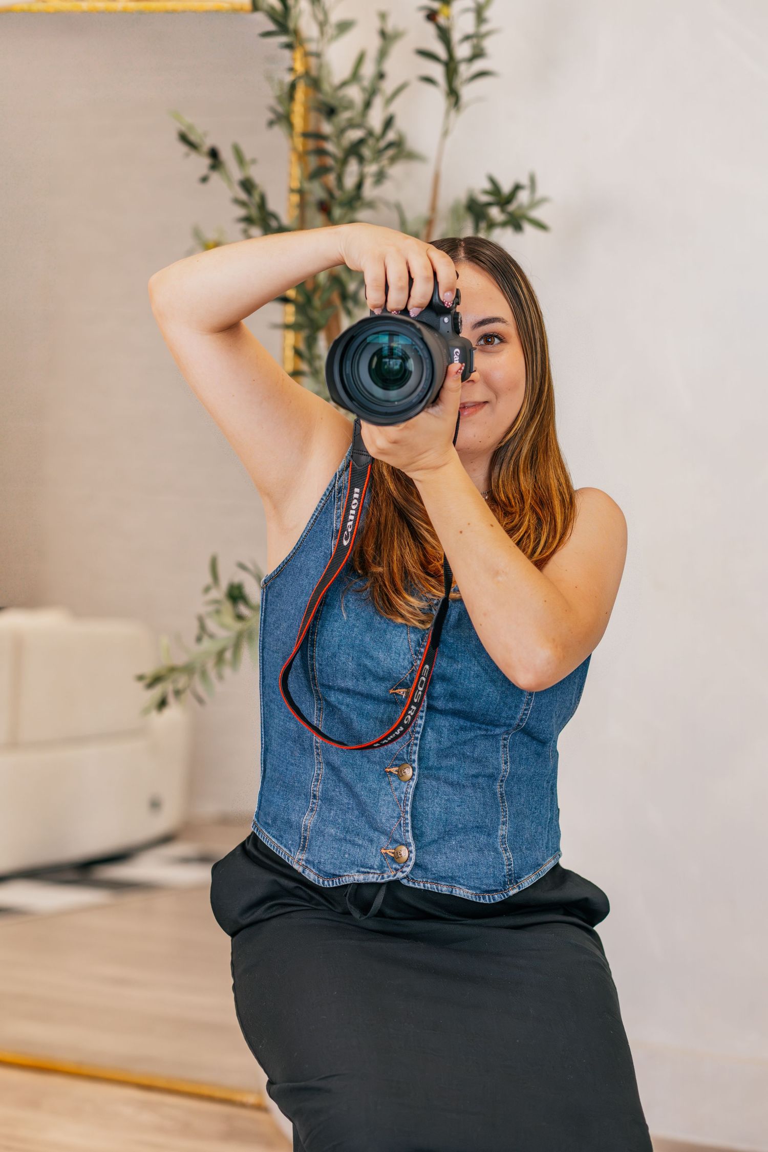 Woman in denim vest holds DSLR camera up to her eye, photographing in a bright modern studio setting.