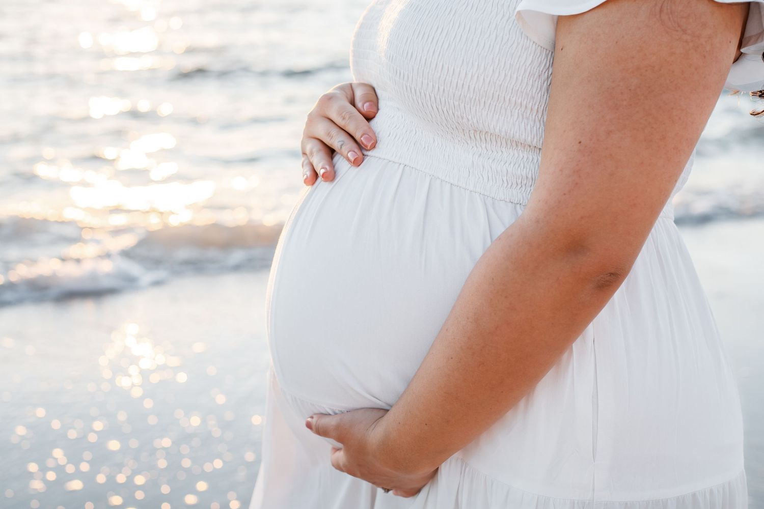 A woman in a white dress cradles her pregnant belly during a beach maternity photoshoot at sunset.
