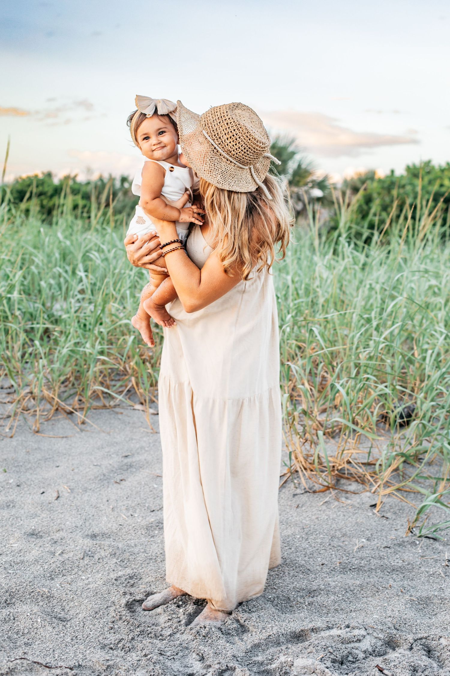 A figure in a flowing beige dress and straw hat embraces a small child on a sandy beach at sunset.
