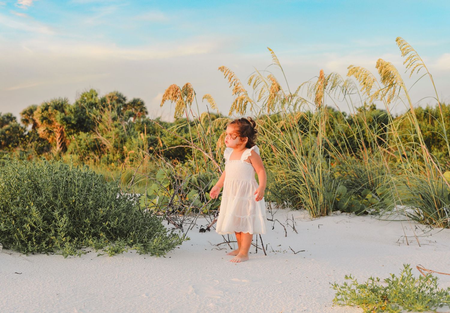 White-dressed figure stands near sea oats during golden hour at a tranquil beach with palm trees in distance.