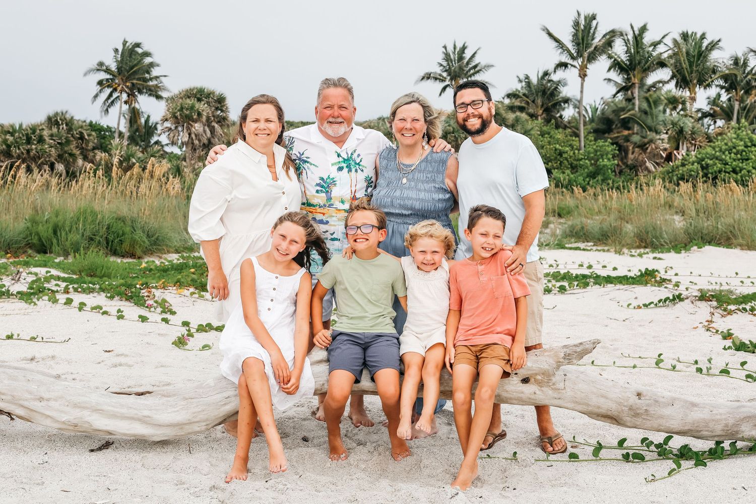 Group enjoying a sunny beach day sitting on driftwood with tropical palm trees and vegetation in the background.