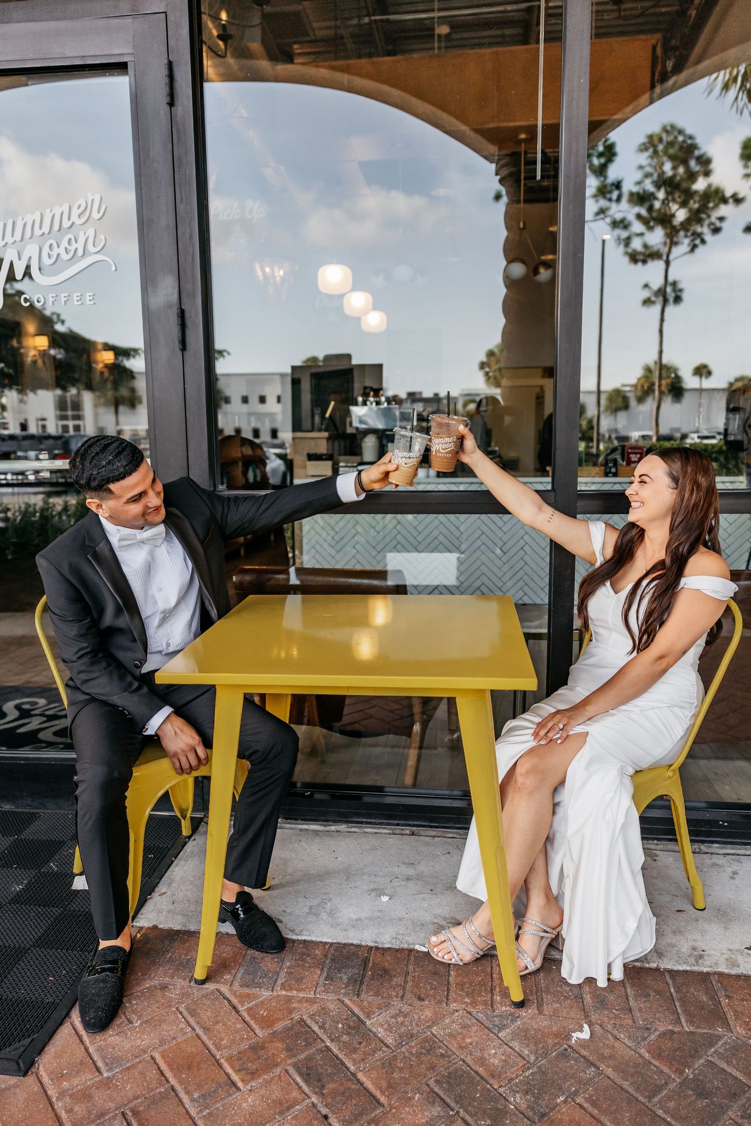 Bride and groom toasting iced coffees at a yellow table outside Summer Moon Coffee shop.