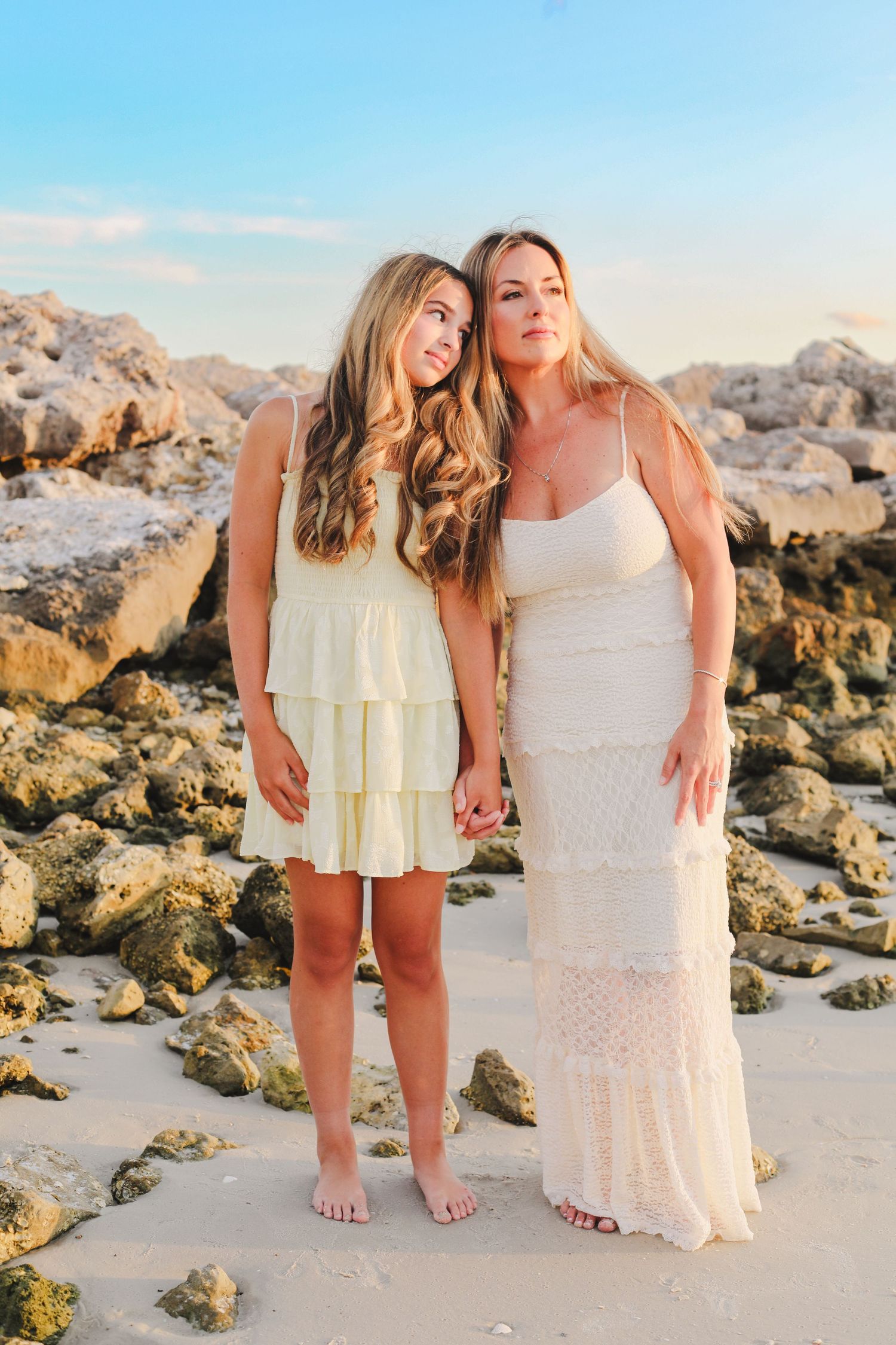 Two women in light dresses pose together on rocky beach at sunset.