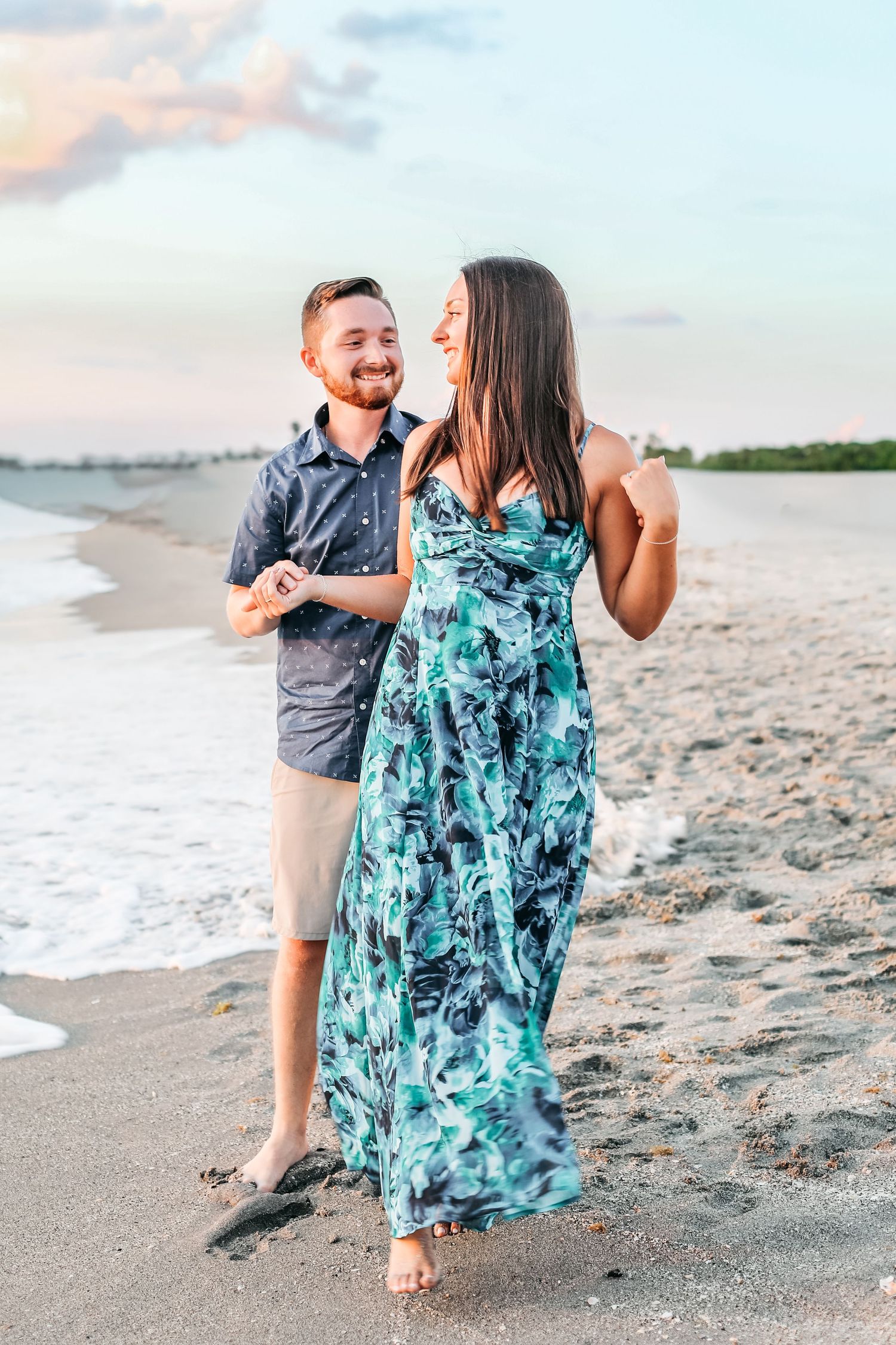 A couple enjoys a romantic stroll along a sandy beach at sunset, with the woman wearing a flowing turquoise dress.