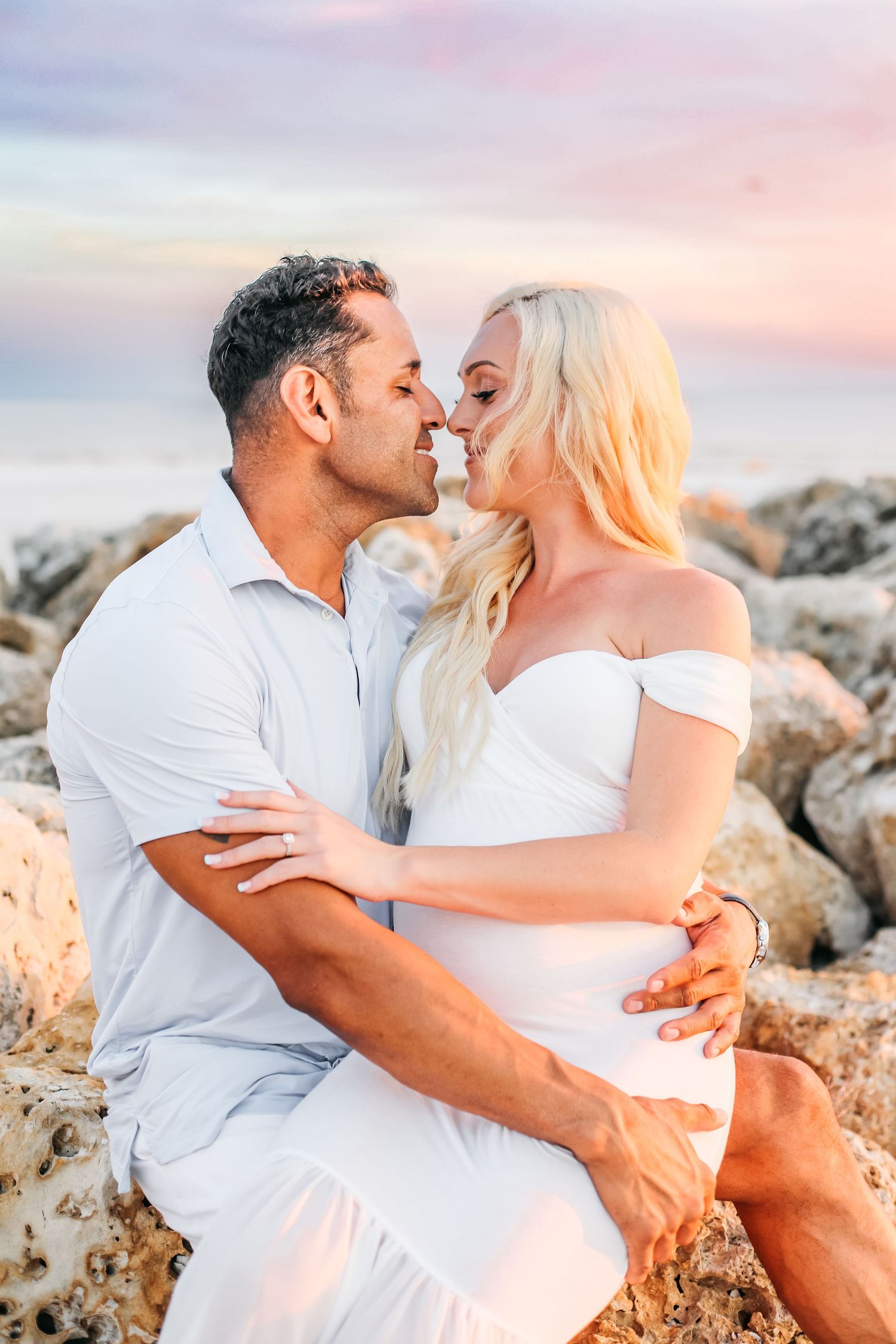 A romantic couple in white outfits embraces on a rocky beach during sunset creating intimate moments.