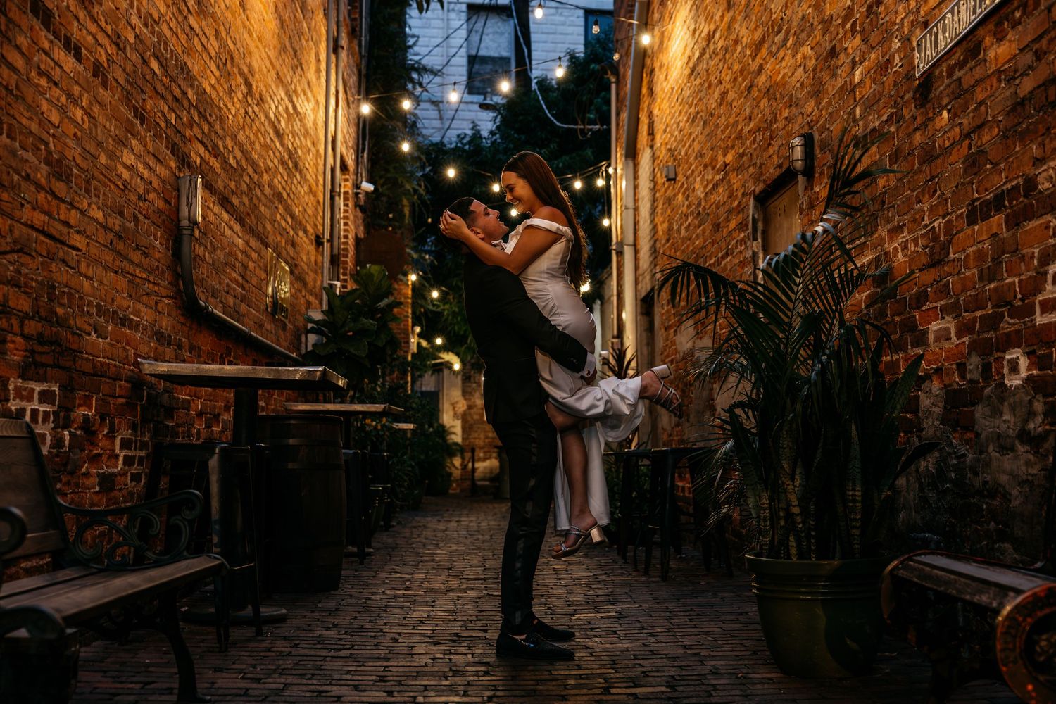 Couple embracing in a romantic brick alley at night, illuminated by warm string lights and lush greenery.