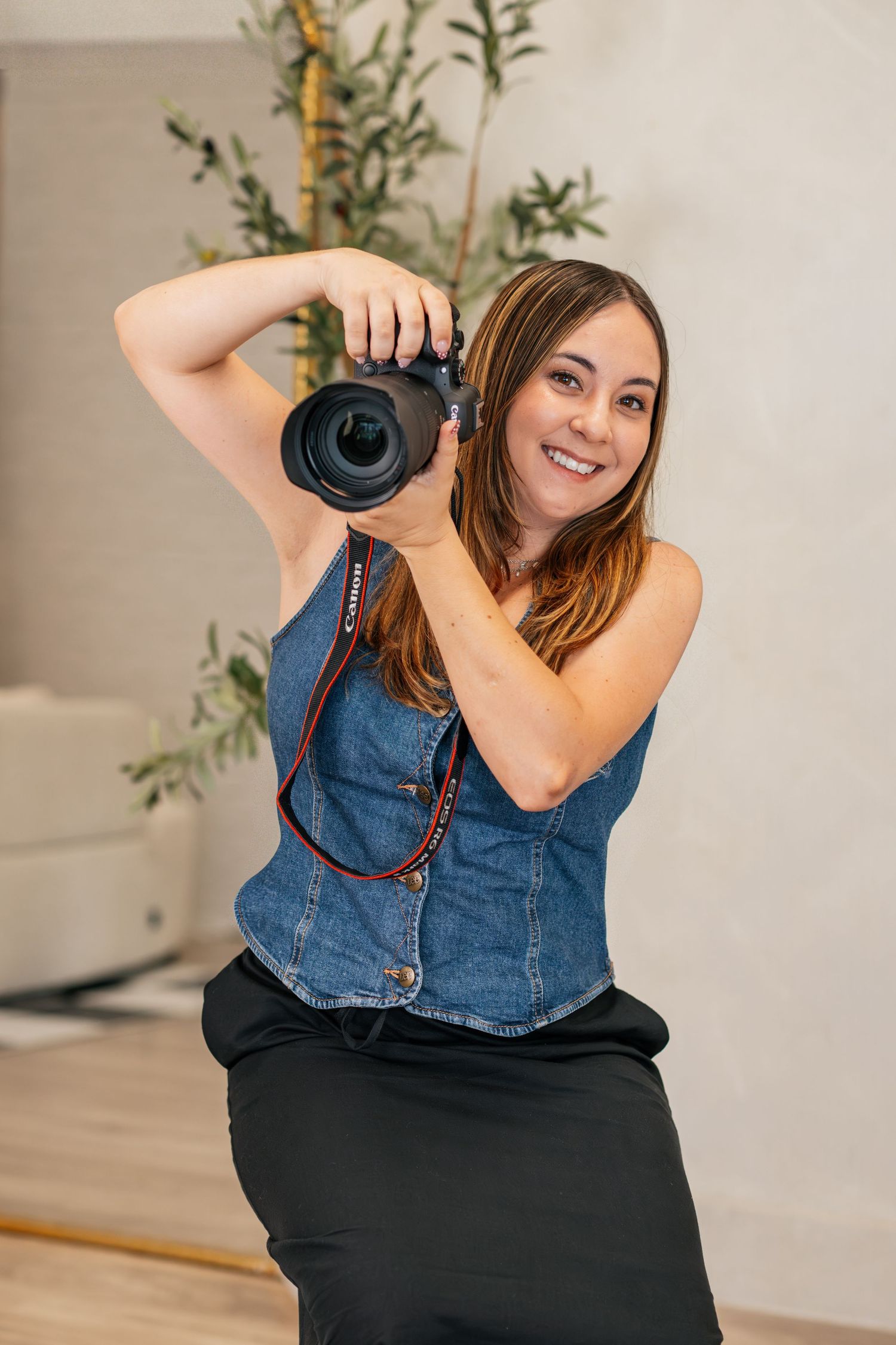 Smiling woman in denim top holding a DSLR camera, posing in a bright modern studio setting.