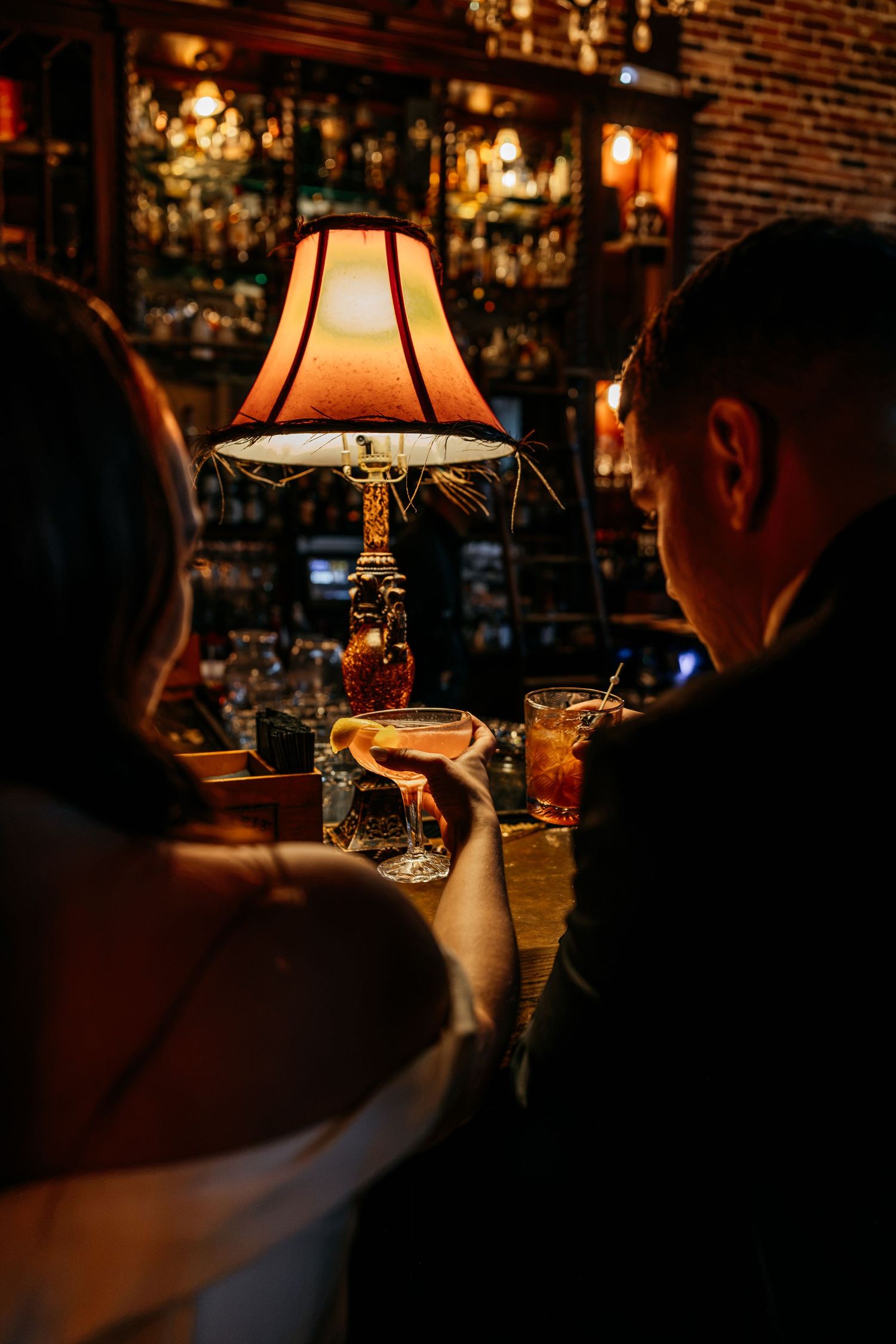 Couple enjoying cocktails at a dimly lit bar, illuminated by a warm vintage table lamp.