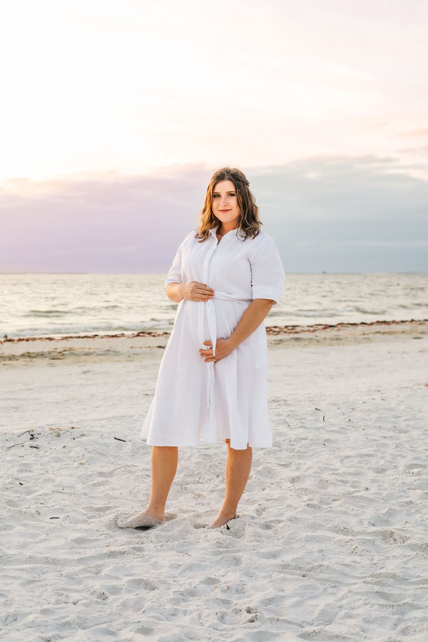 Pregnant woman in white dress cradling baby bump on sandy beach at sunset.