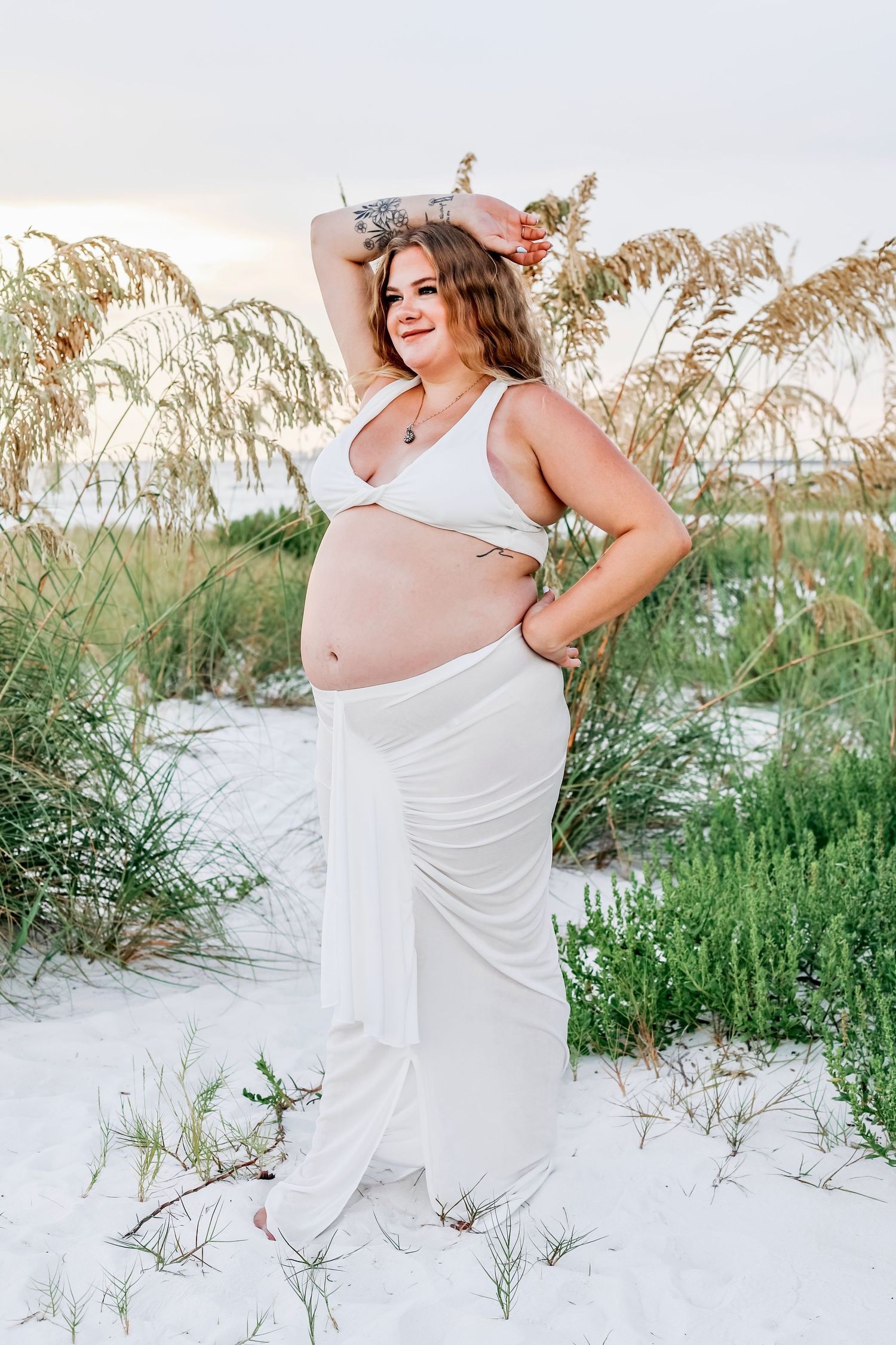 A maternity photoshoot on white sand dunes with sea oats at sunset.