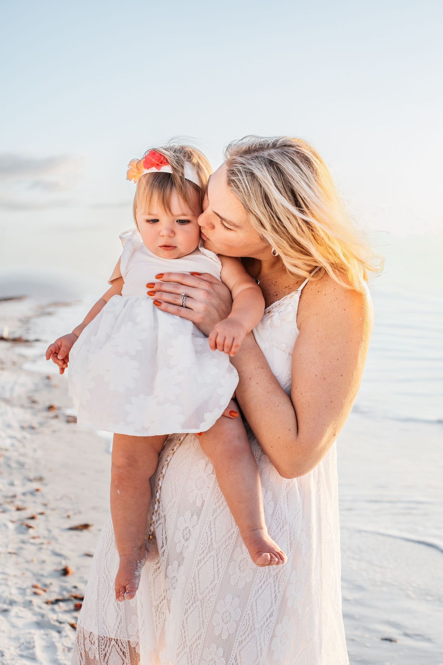 A tender beach moment captured at sunset with white lace dresses against the sandy shore and ocean backdrop.