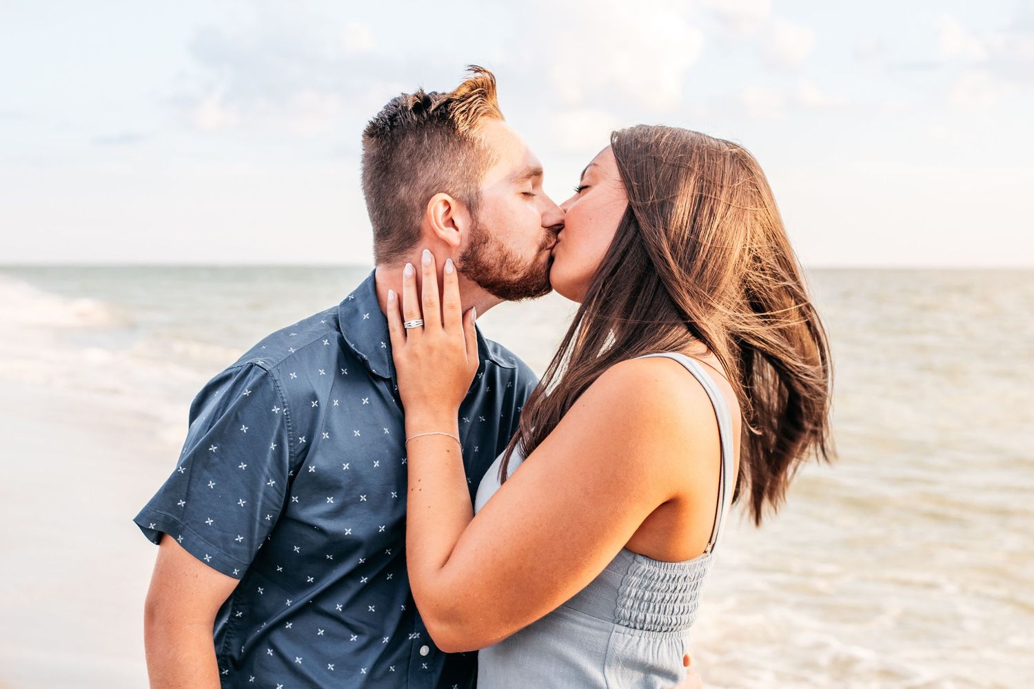 A couple shares a romantic kiss on the beach with the ocean and cloudy sky in the background.
