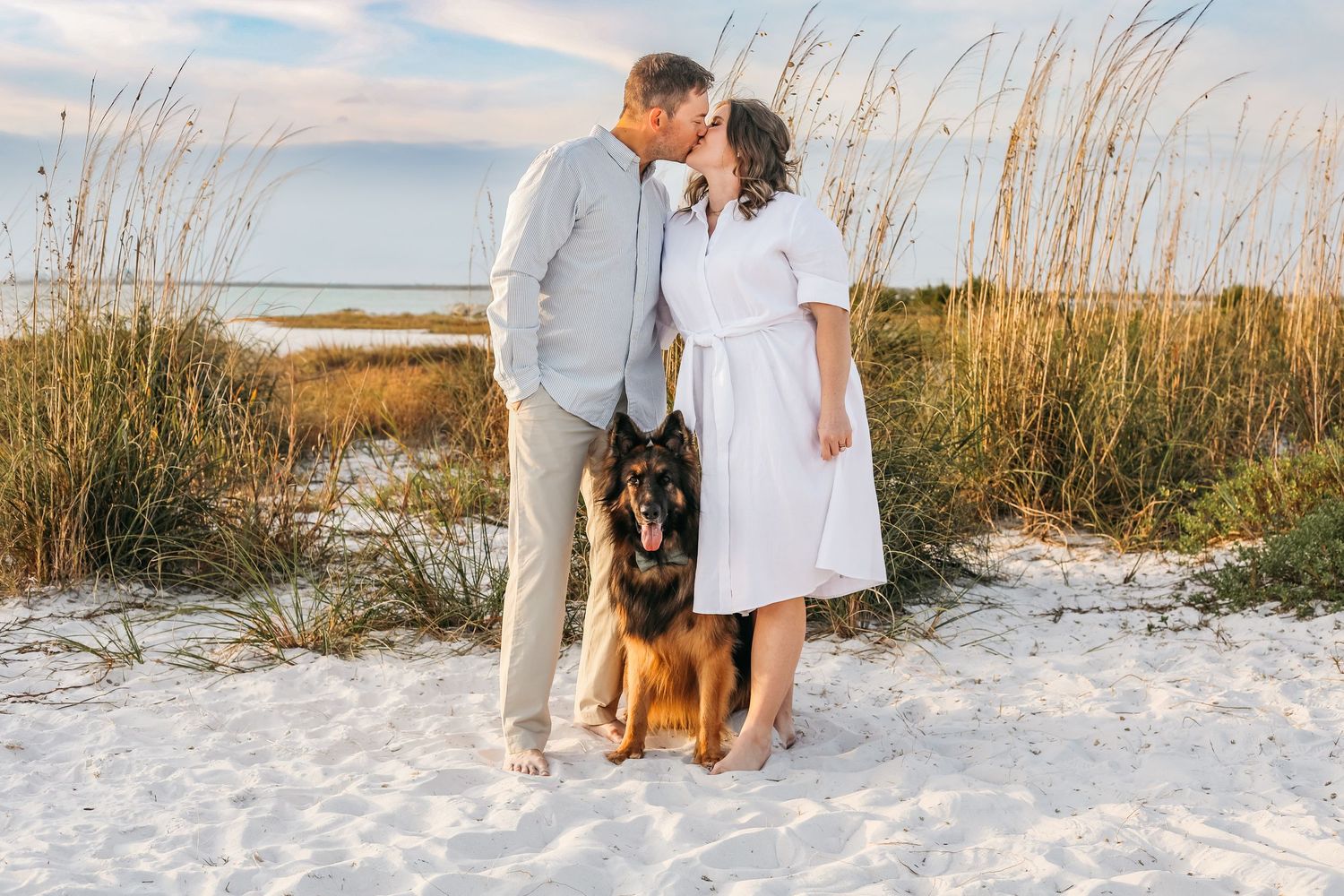 Couple kissing on white sand beach with German Shepherd dog, sea oats in background at golden hour.