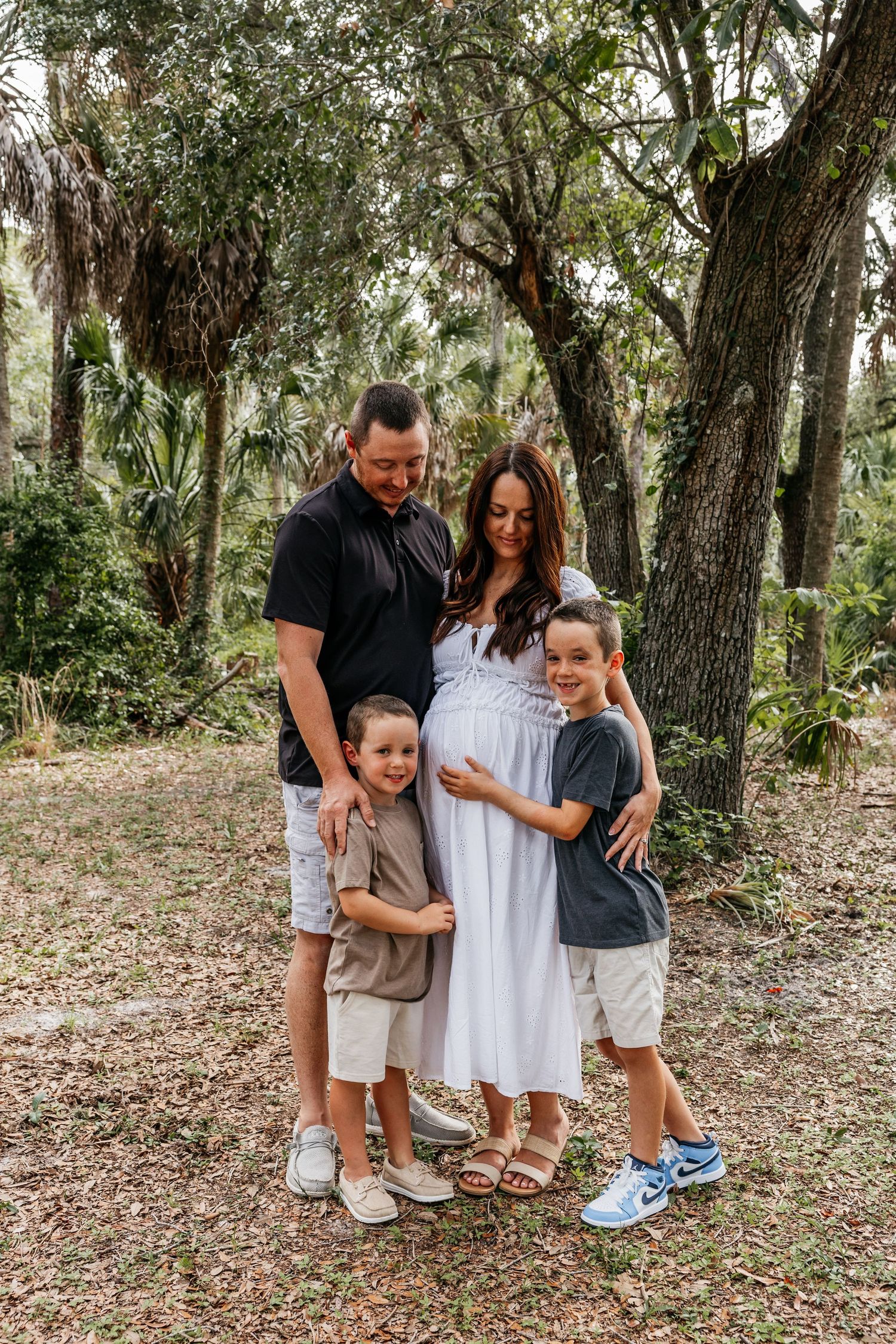 Pregnant woman in white dress poses with husband and two young sons in a lush outdoor setting for a maternity photo.