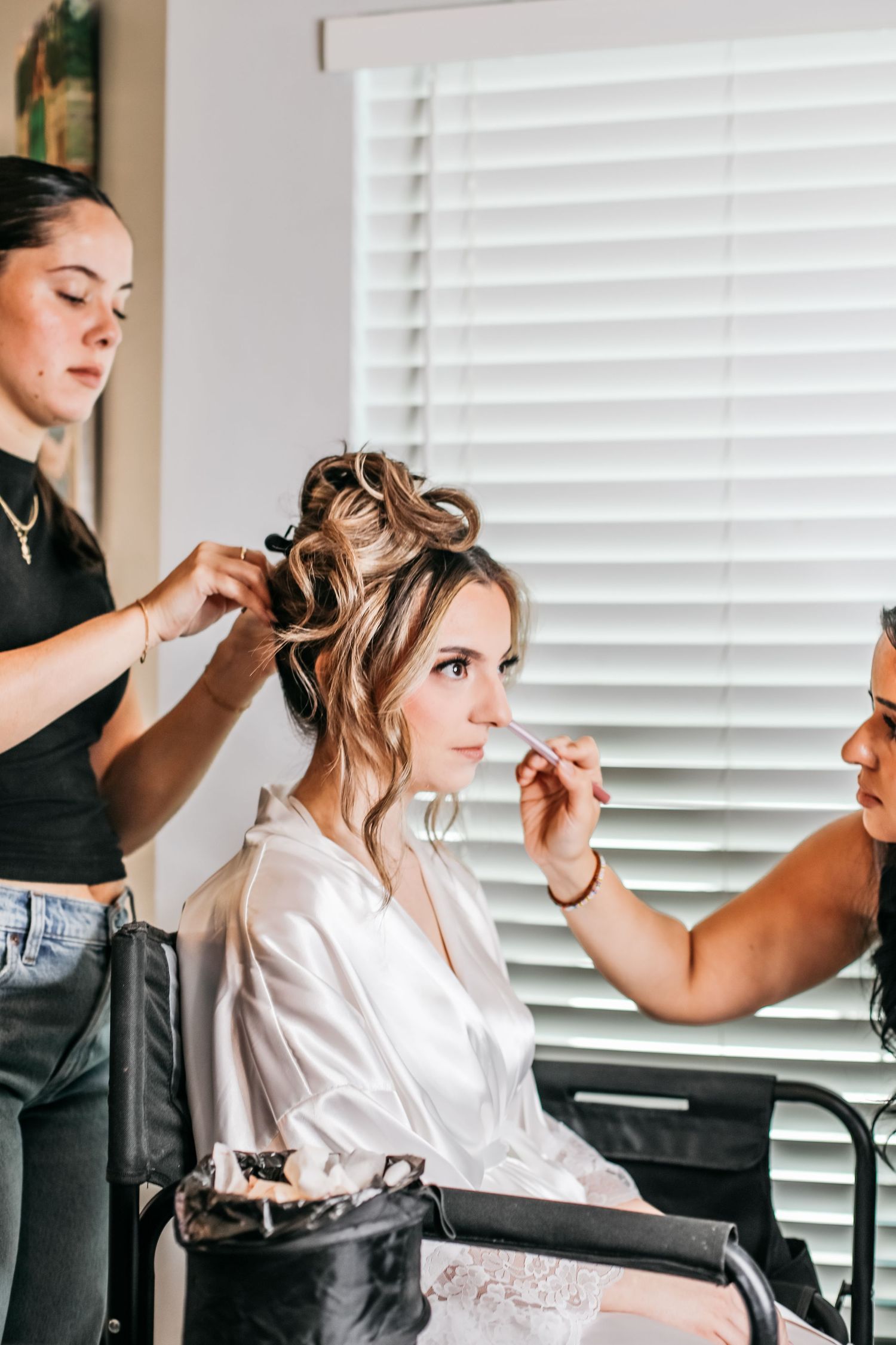 Two beauty artists style a bride's hair and apply makeup as she sits in a salon chair by a window.