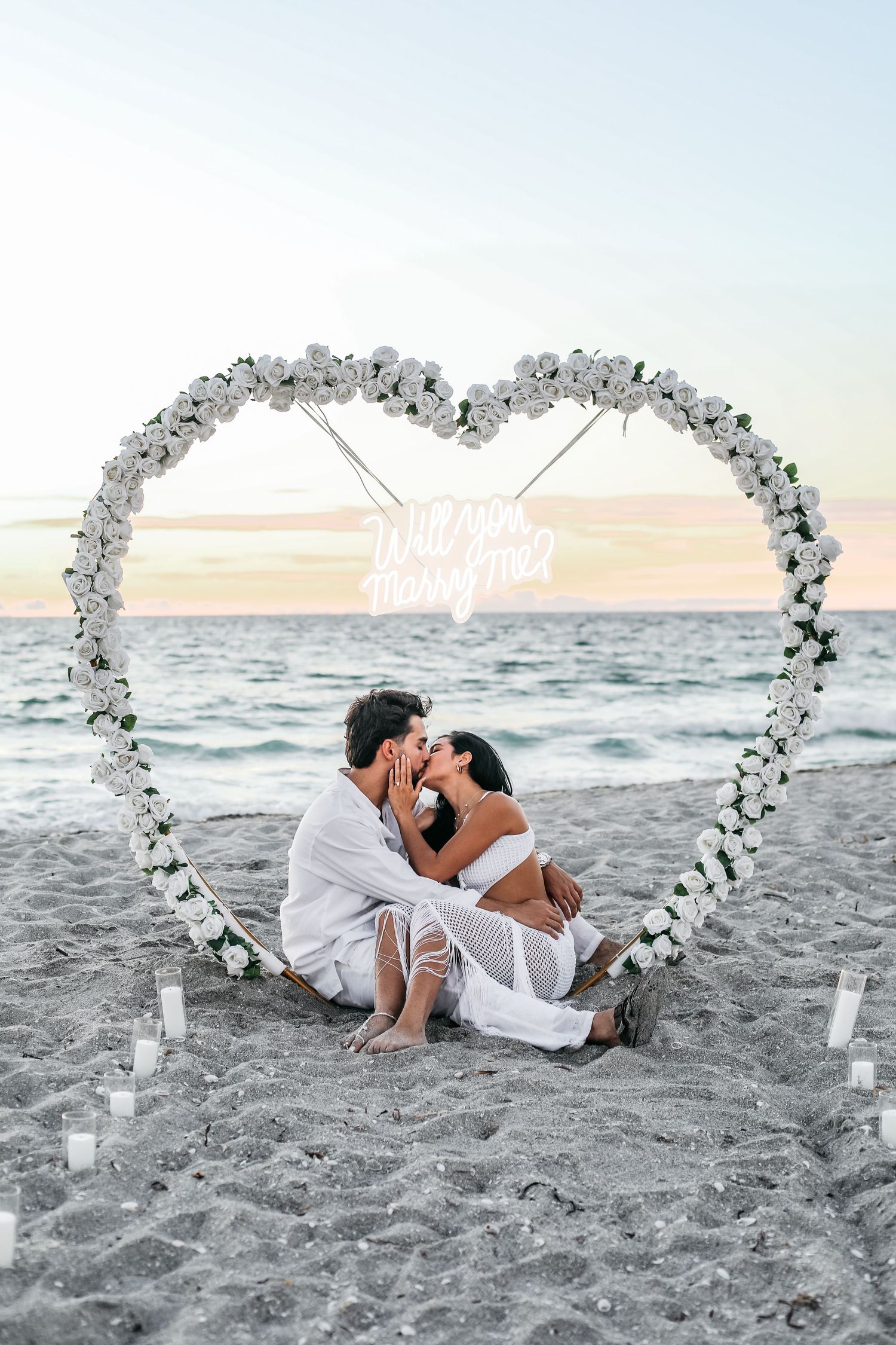 A romantic beach scene with a heart-shaped floral arch and neon sign framing a couple sitting on the sand at sunset.