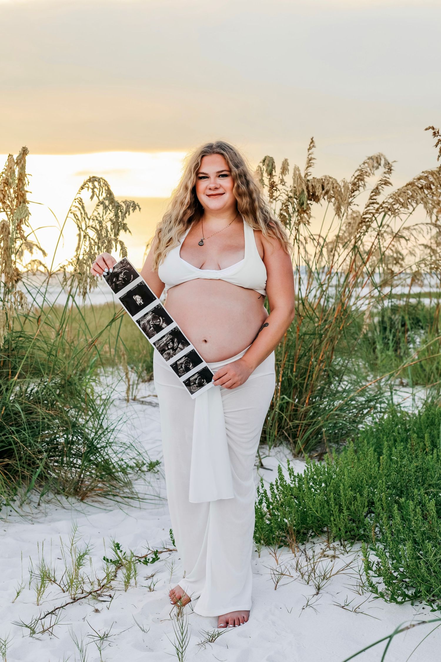 Woman in white dress poses with ultrasound photos on sandy beach at sunset.