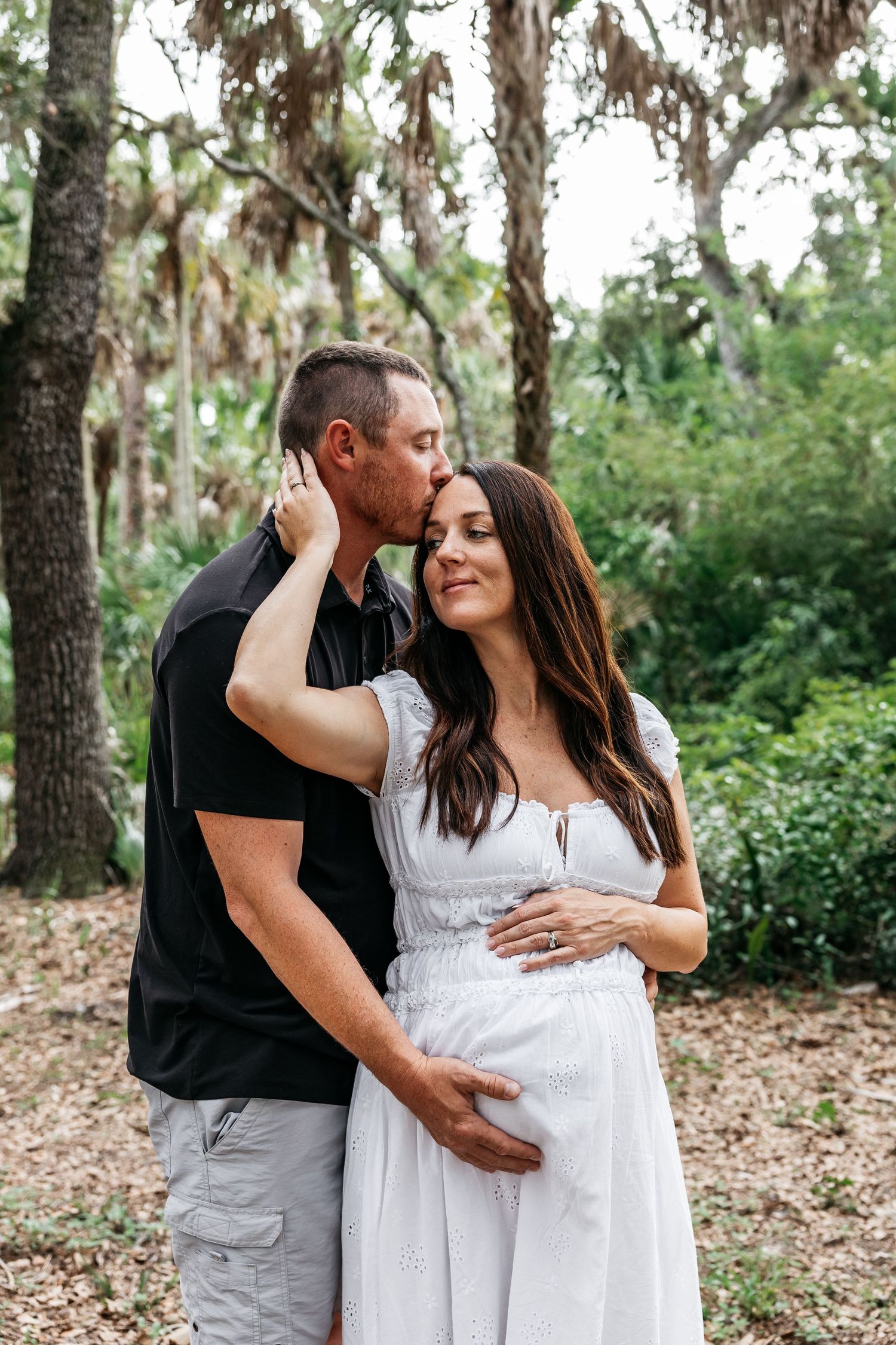 A man kisses a pregnant woman in a white dress on the cheek outdoors, surrounded by lush green trees.