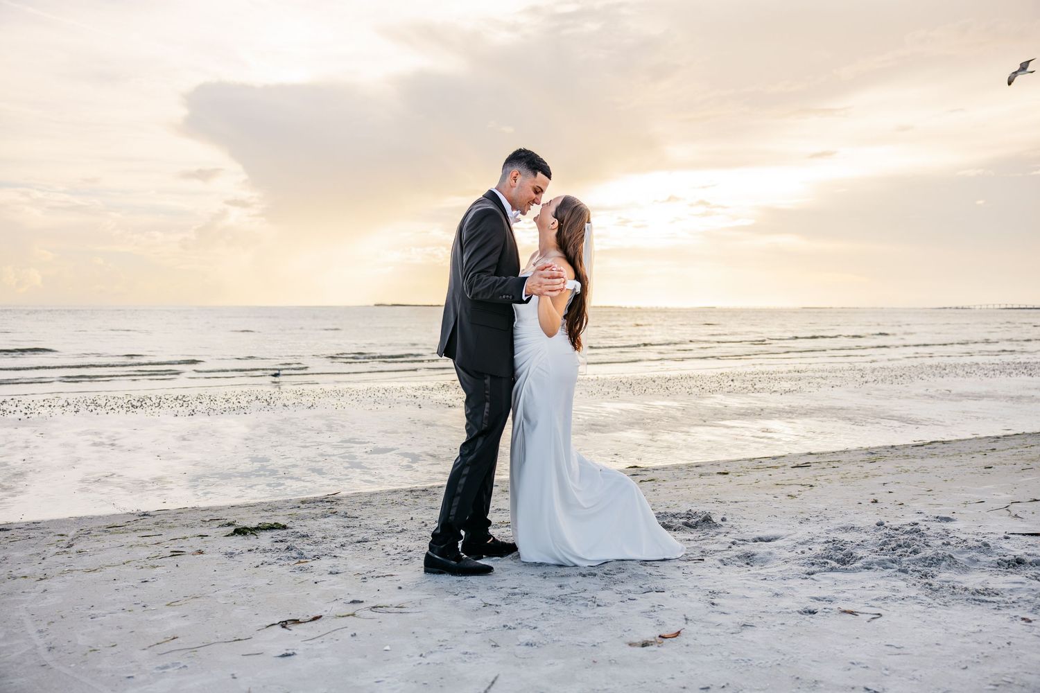 Couple sharing a romantic kiss on the beach at sunset, bride in white gown and groom in dark suit.