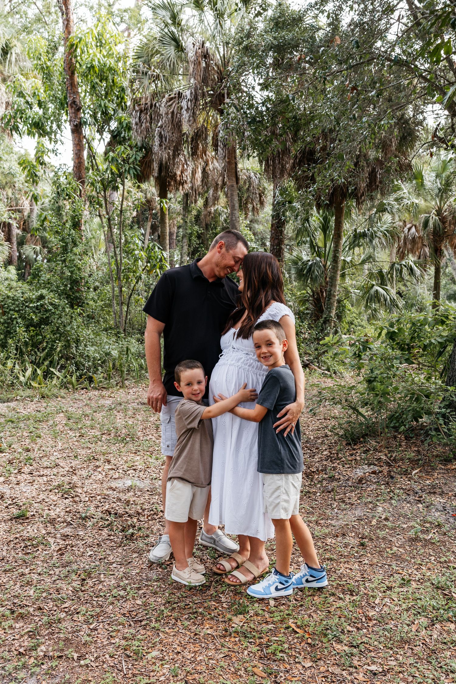 Pregnant woman in white dress with husband and two boys hugging her baby bump outdoors near palm trees.