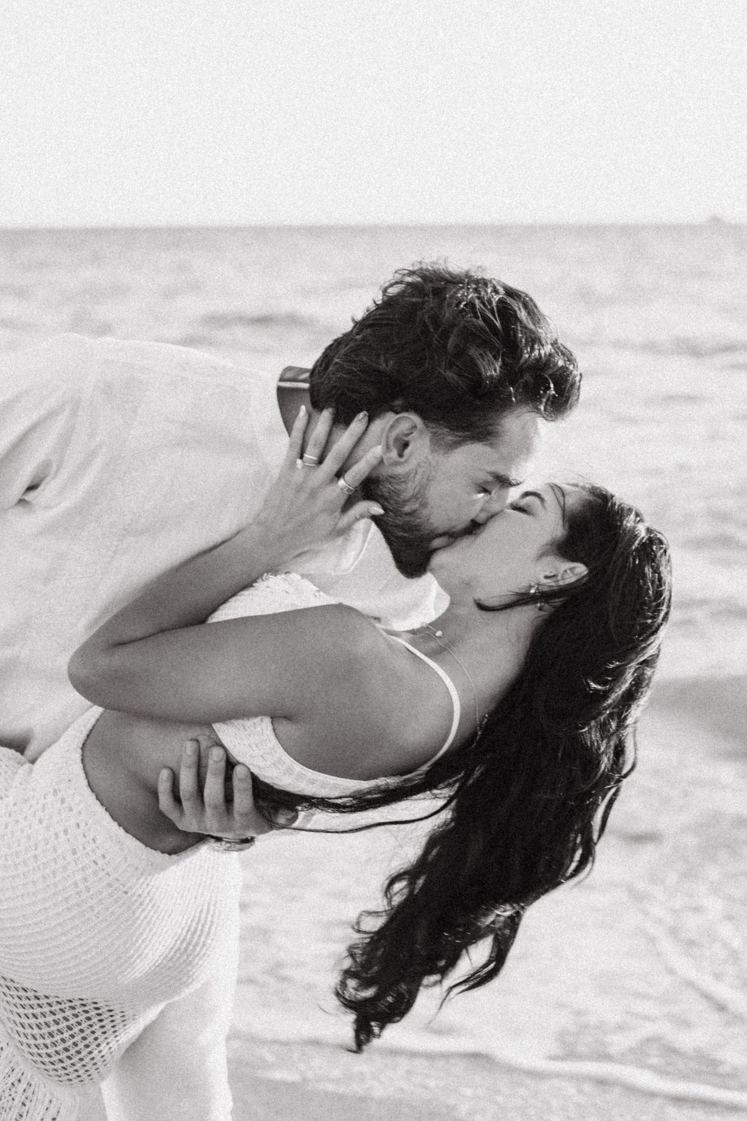 A romantic black and white photo of a couple embracing and kissing on the beach with ocean waves in the background.