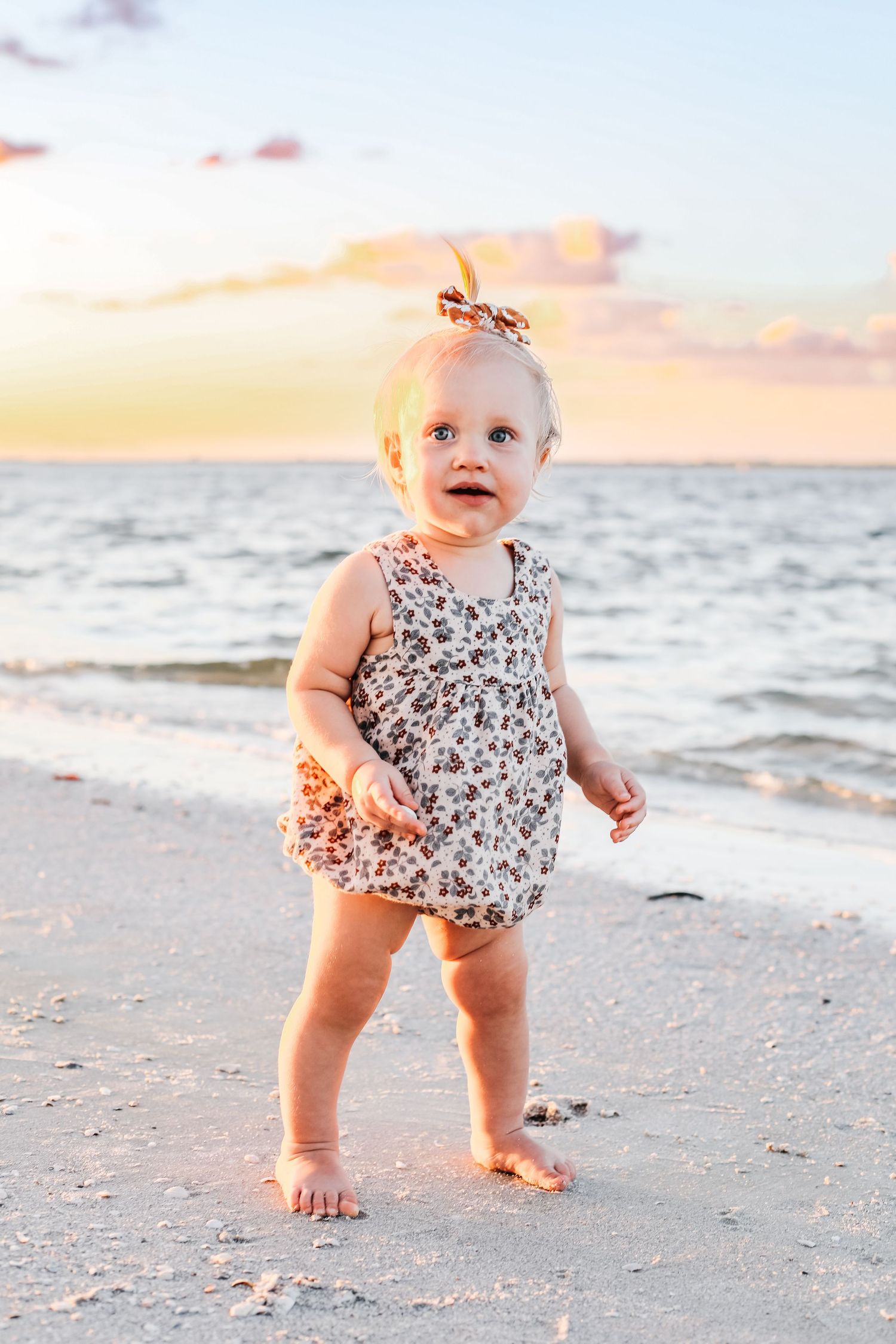 Baby in floral romper exploring sandy beach at sunset with golden light.