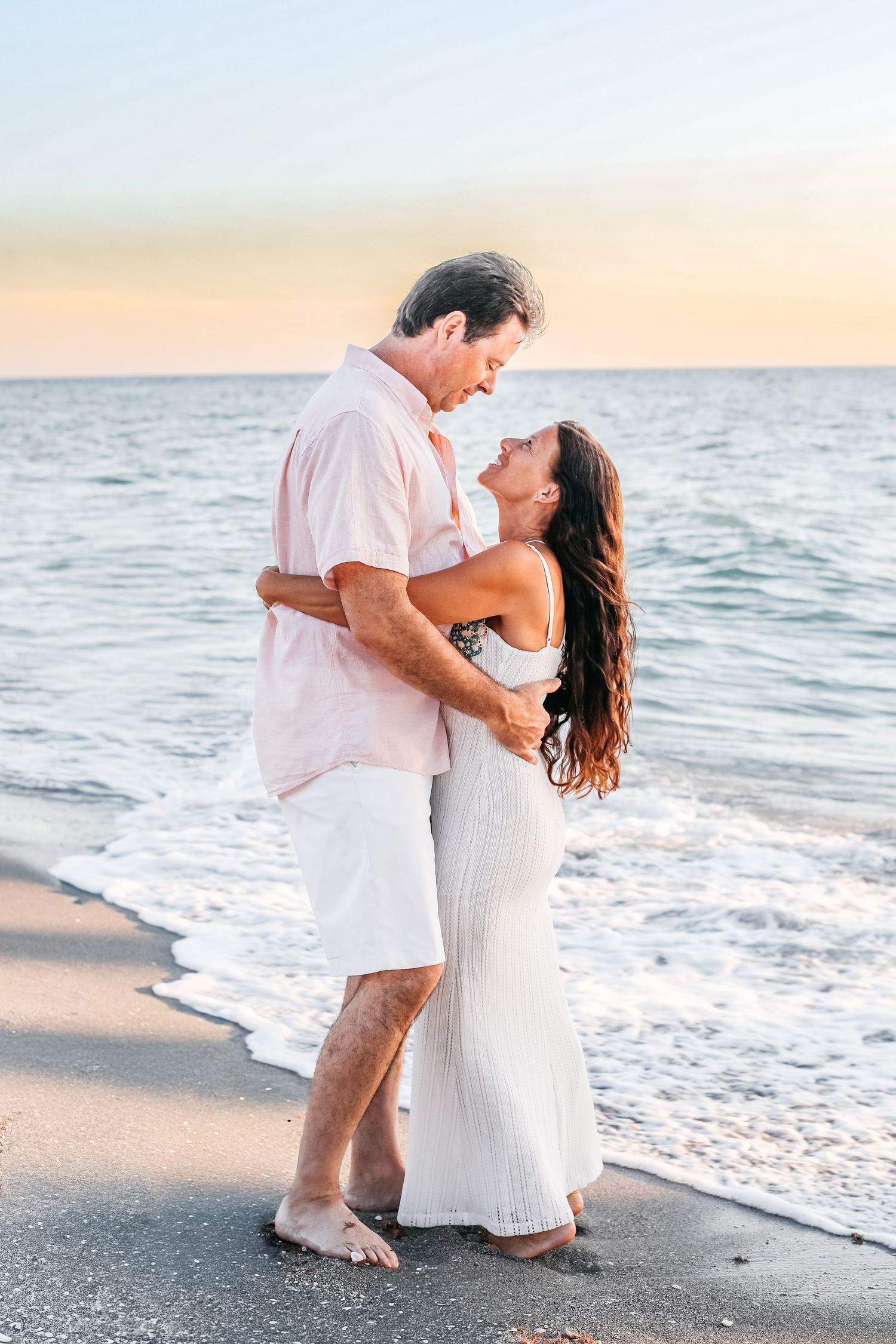 A romantic couple in white attire embraces on a sandy beach at sunset with ocean waves in the background.