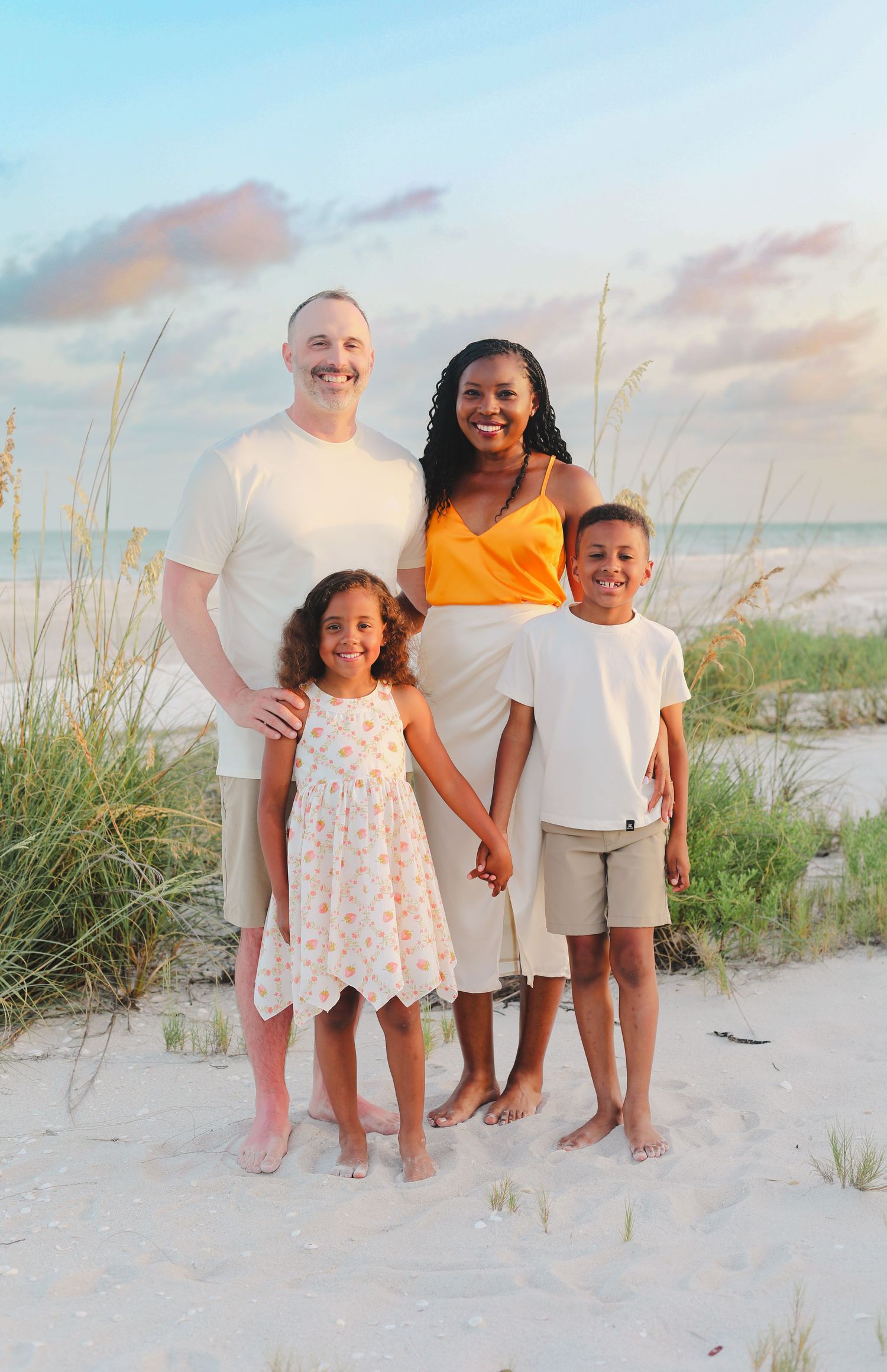 Family in white and light colored outfits posing together on white sandy beach with sea grass.