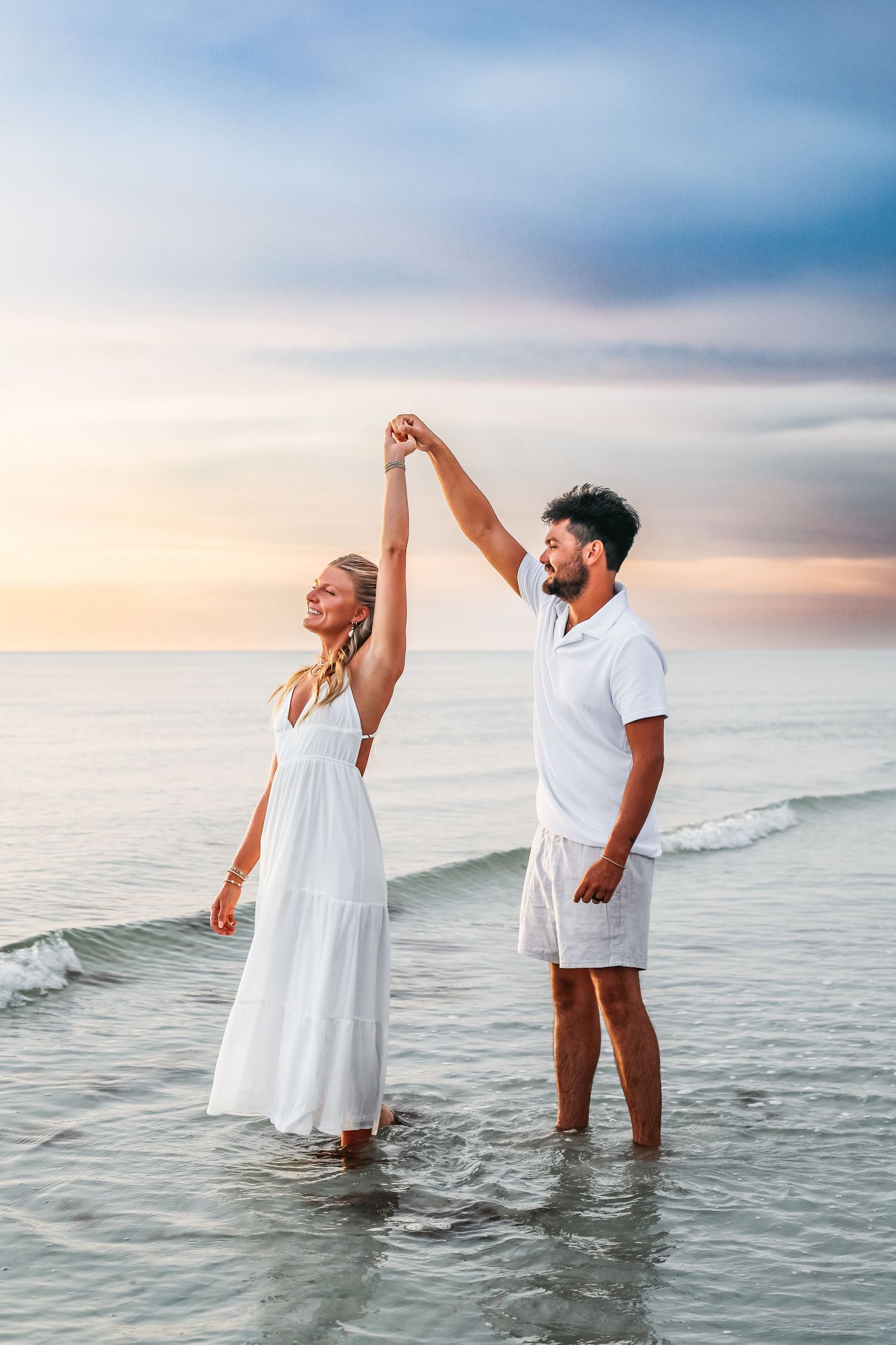 Couple in white clothing playing in ocean waves during sunset photoshoot.