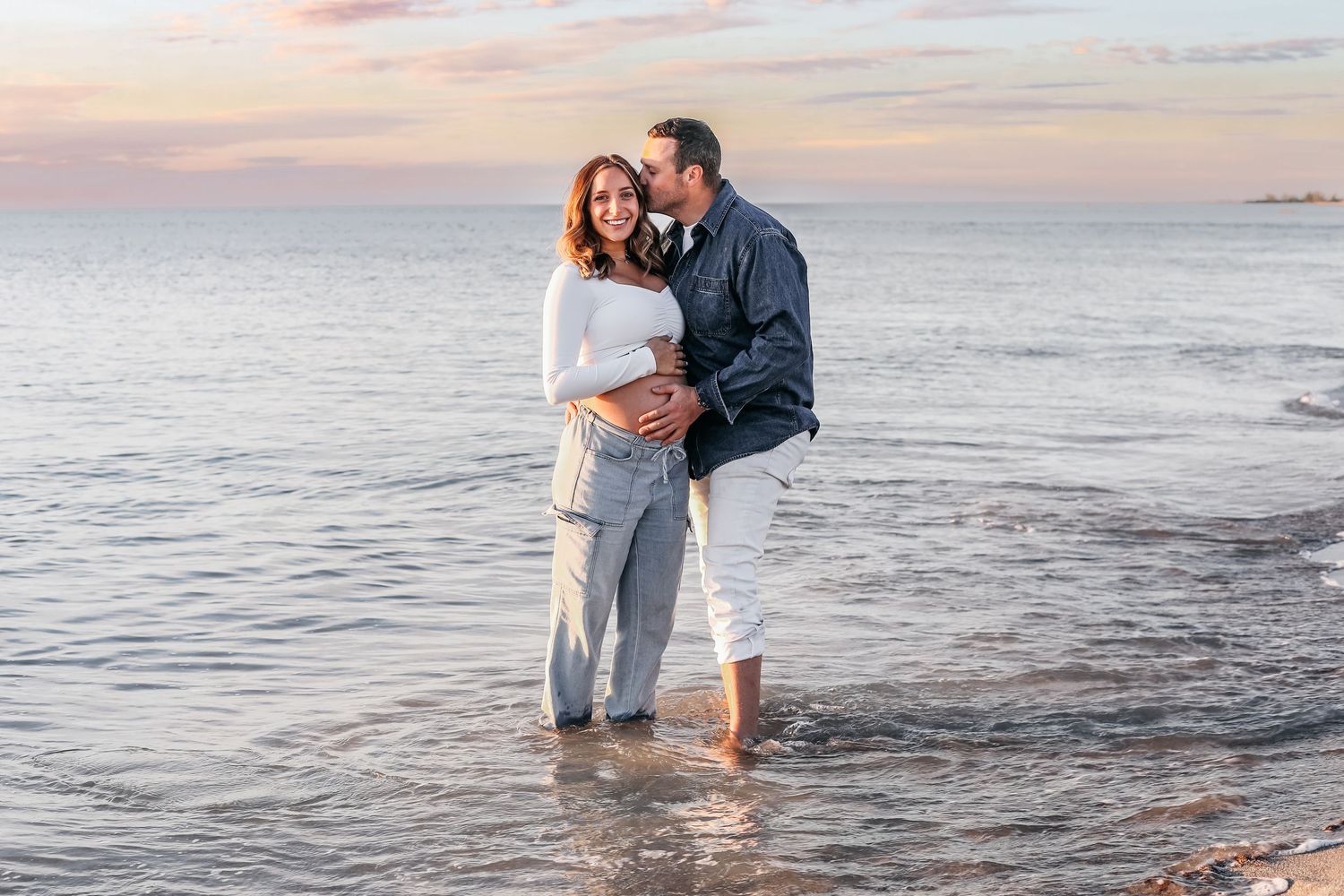 Expectant parents share tender moments while standing in shallow ocean waters at dusk.