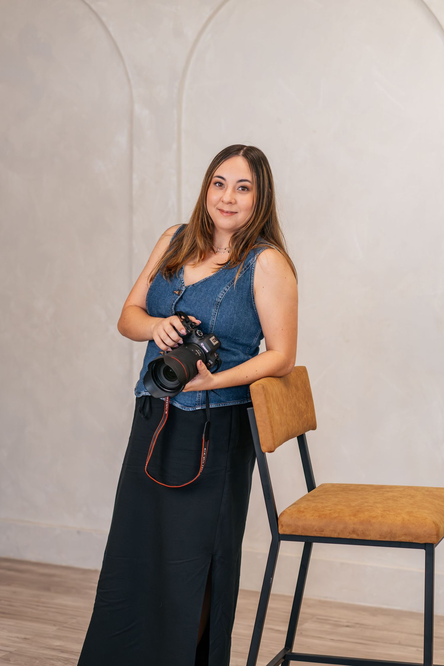 Woman photographer in denim top and black skirt holding DSLR camera, posing beside a wooden chair in a bright studio.