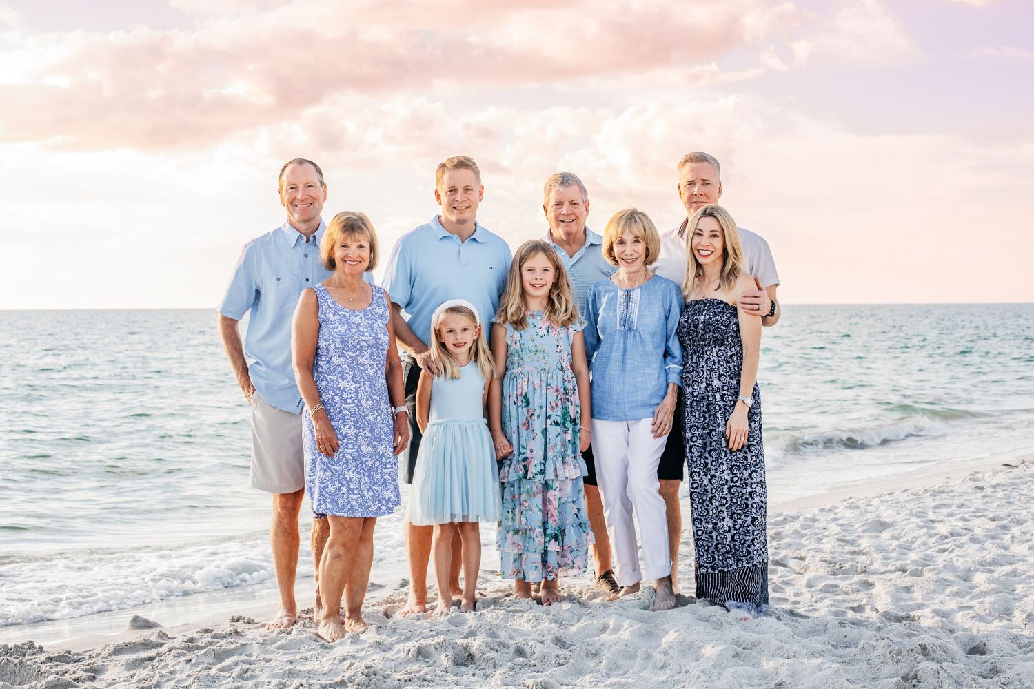 Family of eight posing together on a sandy beach at sunset with pastel skies and ocean waves in the background.