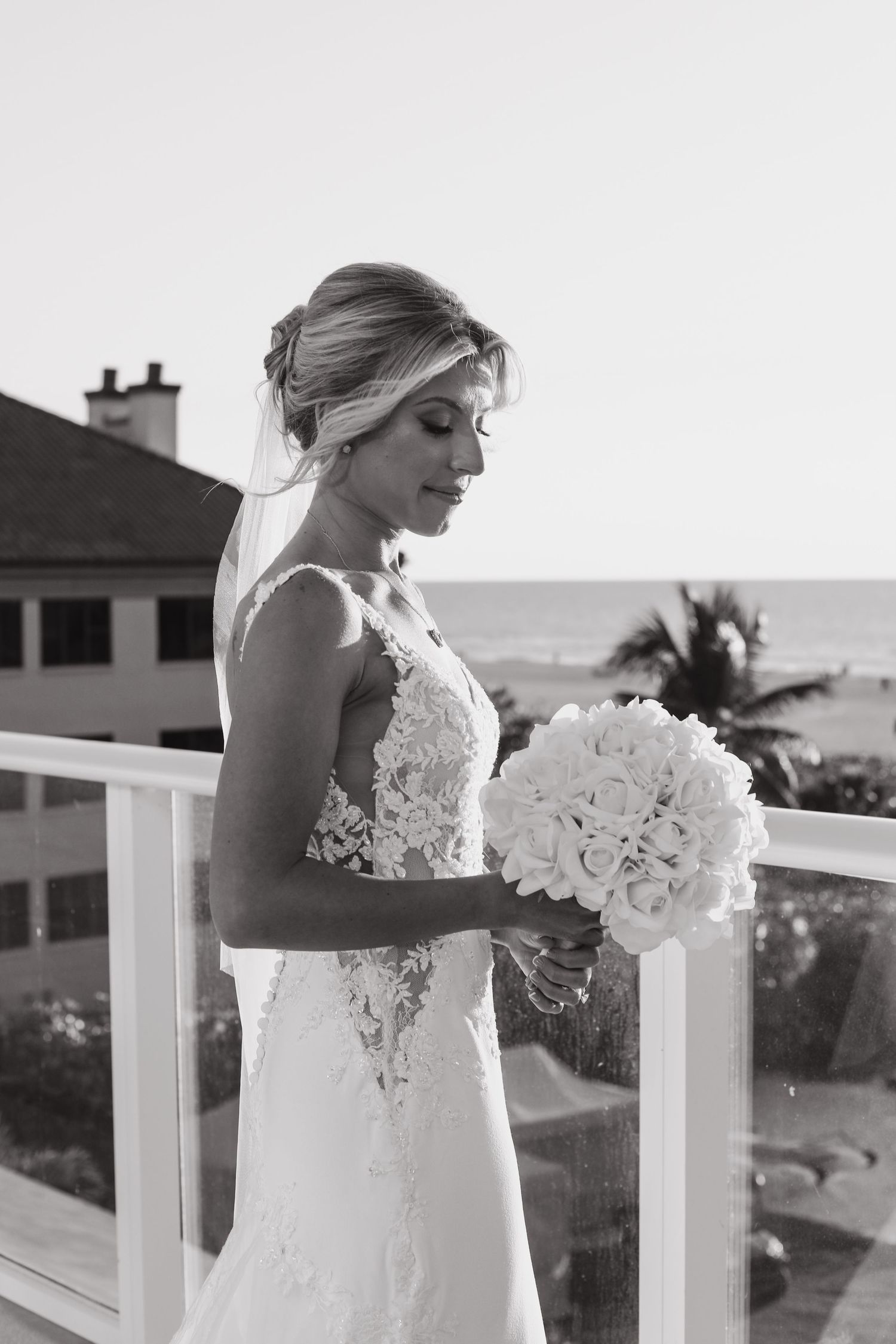 Bride in lace wedding gown holds white rose bouquet on balcony with ocean view, black and white photo.