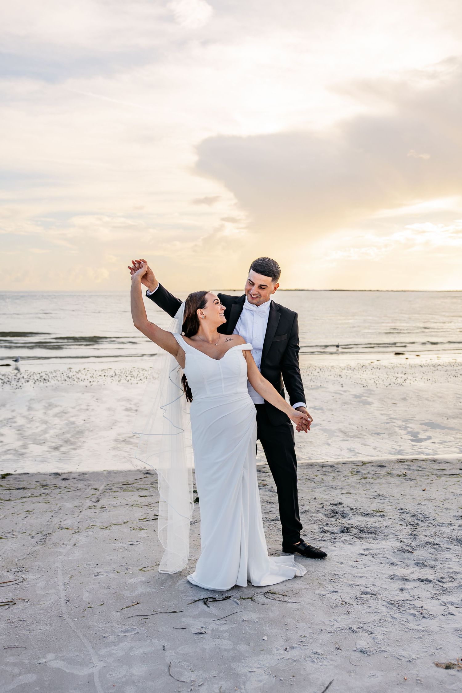Bride and groom dancing on beach at sunset, bride in white gown and groom in black tuxedo.