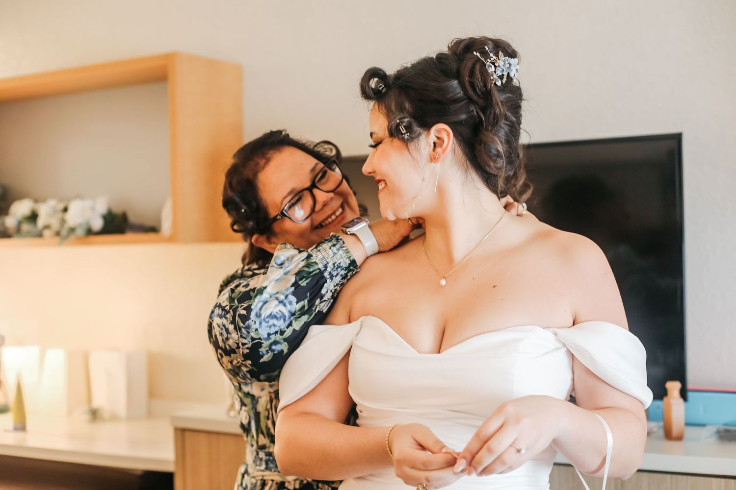 Bride in white off-shoulder gown shares tender moment with woman in floral dress during wedding preparations.
