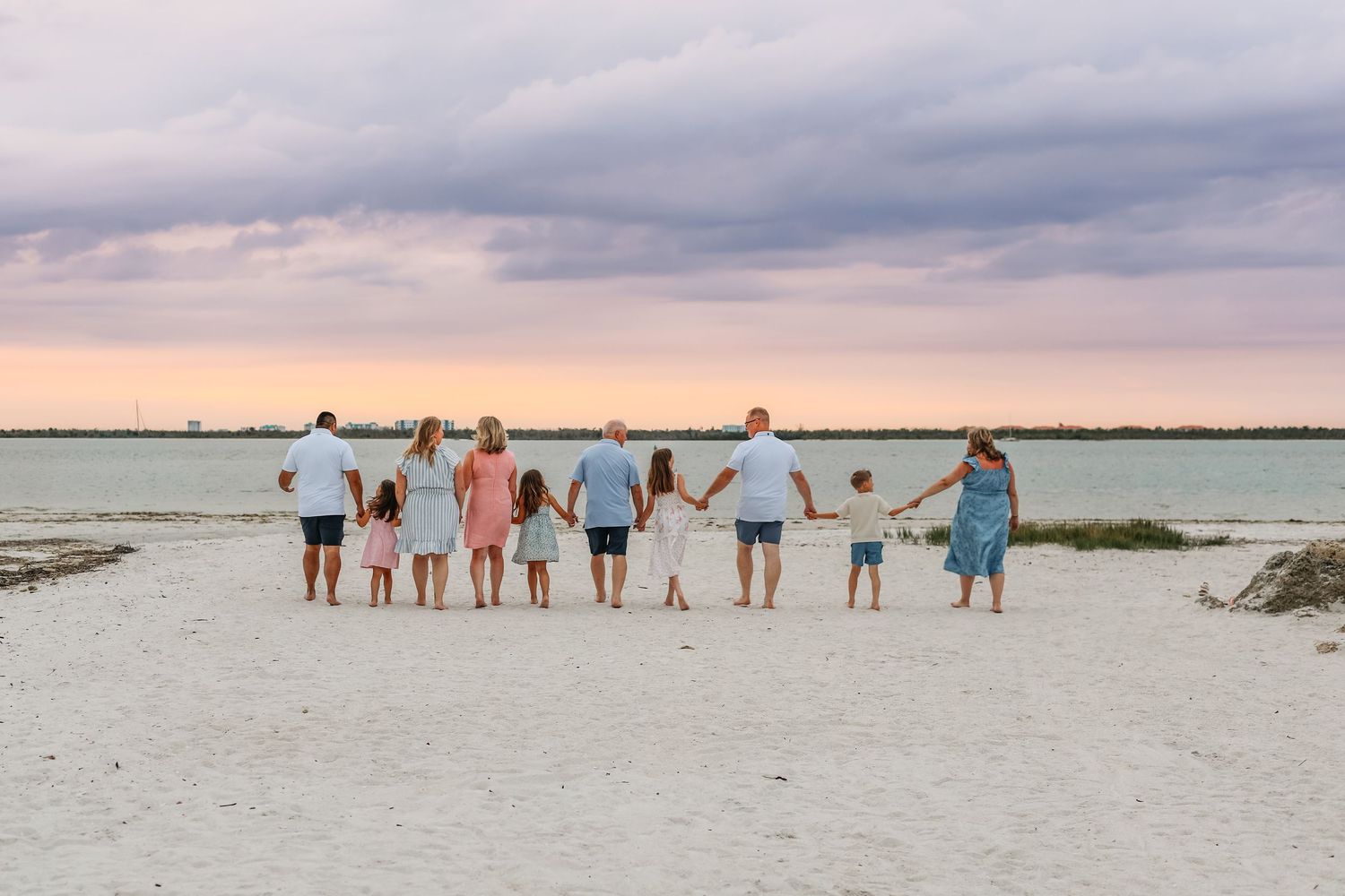 Family holding hands on a sandy beach at sunset with colorful pink and purple sky over calm ocean waters.