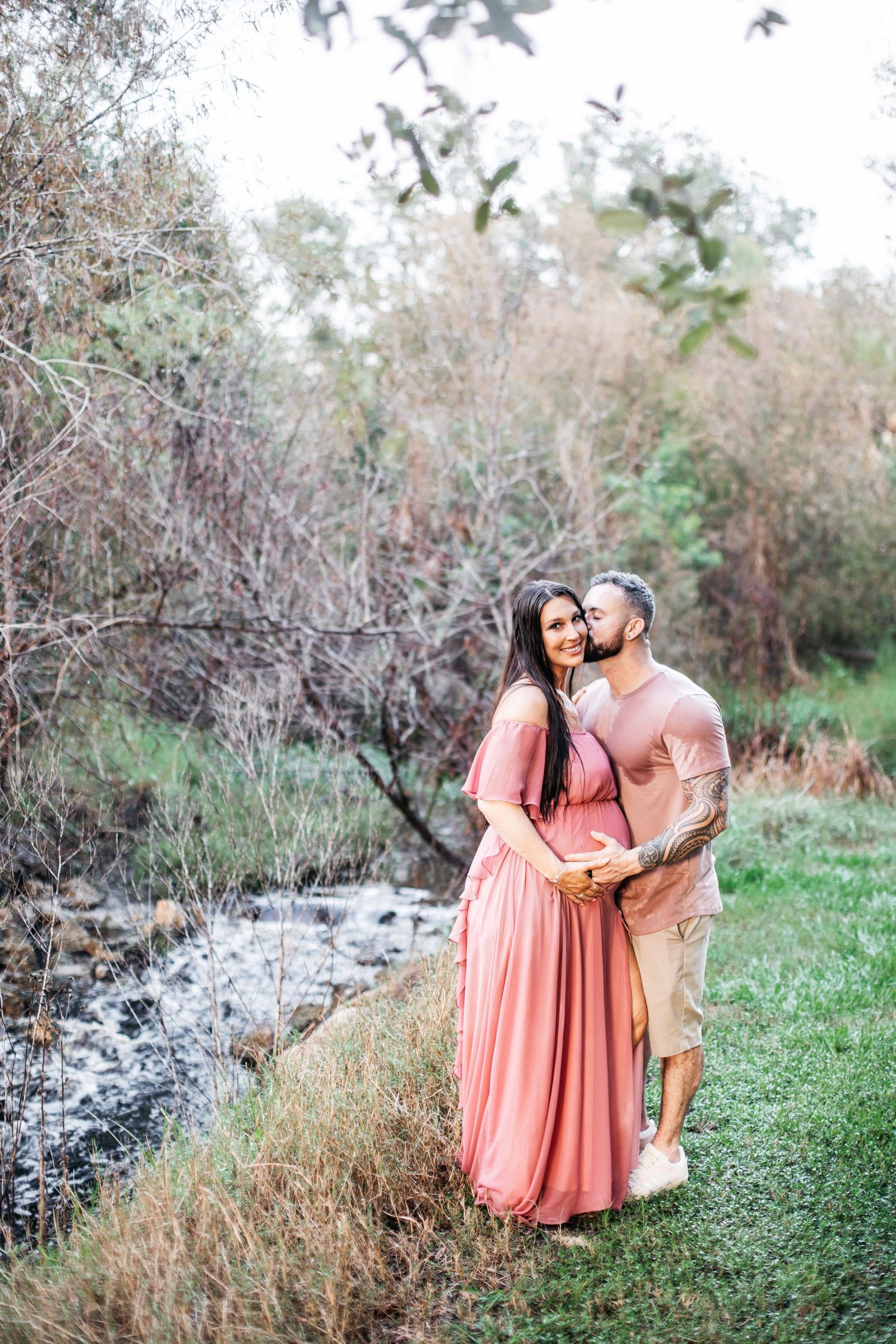 A couple shares an intimate moment together in a natural outdoor setting with tall grass and trees.