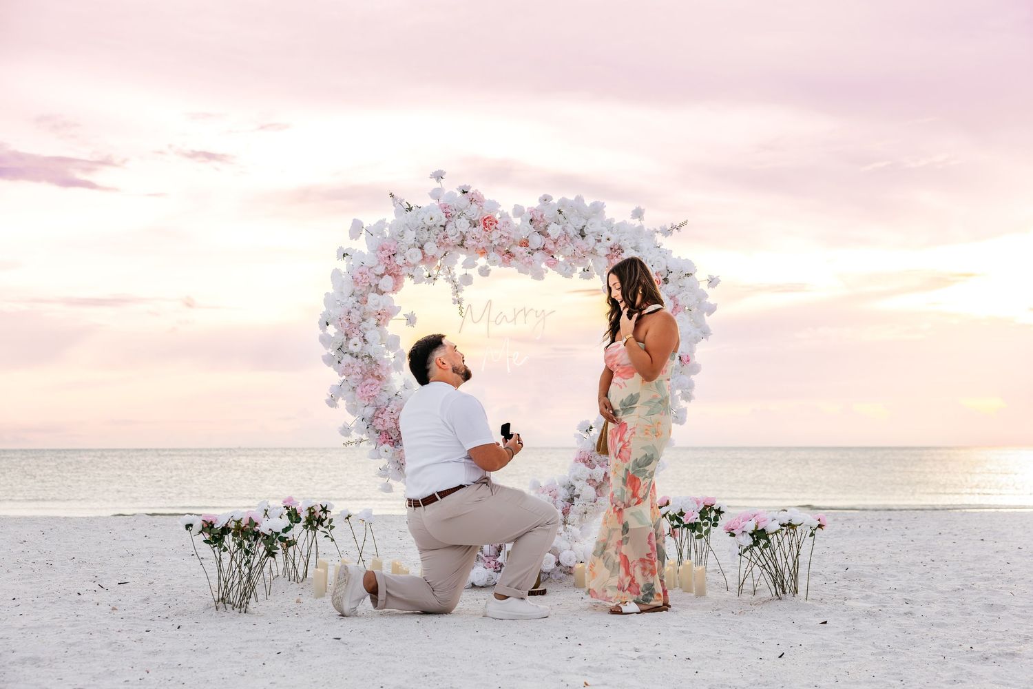 Man proposes to woman on beach at sunset with heart-shaped floral arch and flowers on sand.