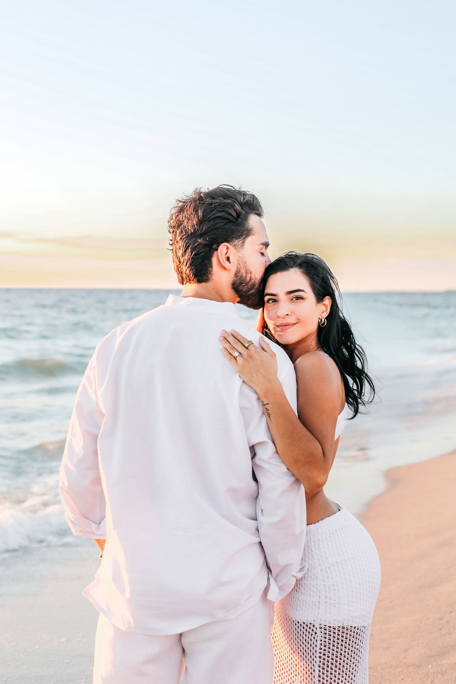 A romantic couple embraces on a sandy beach at sunset while wearing white outfits.