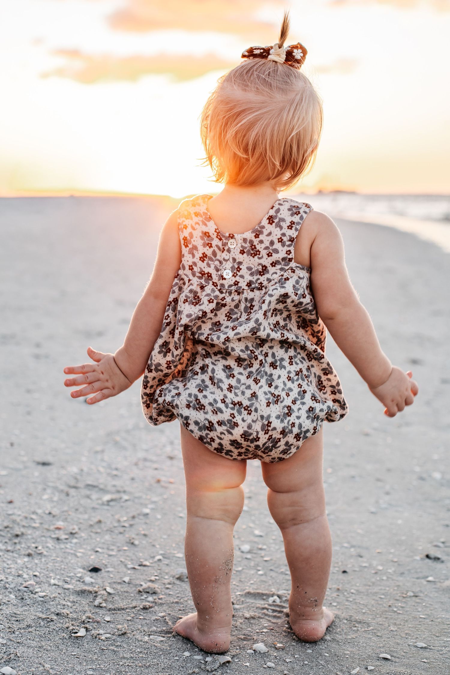 A toddler in a floral romper stands on the beach at sunset with golden light illuminating their back.