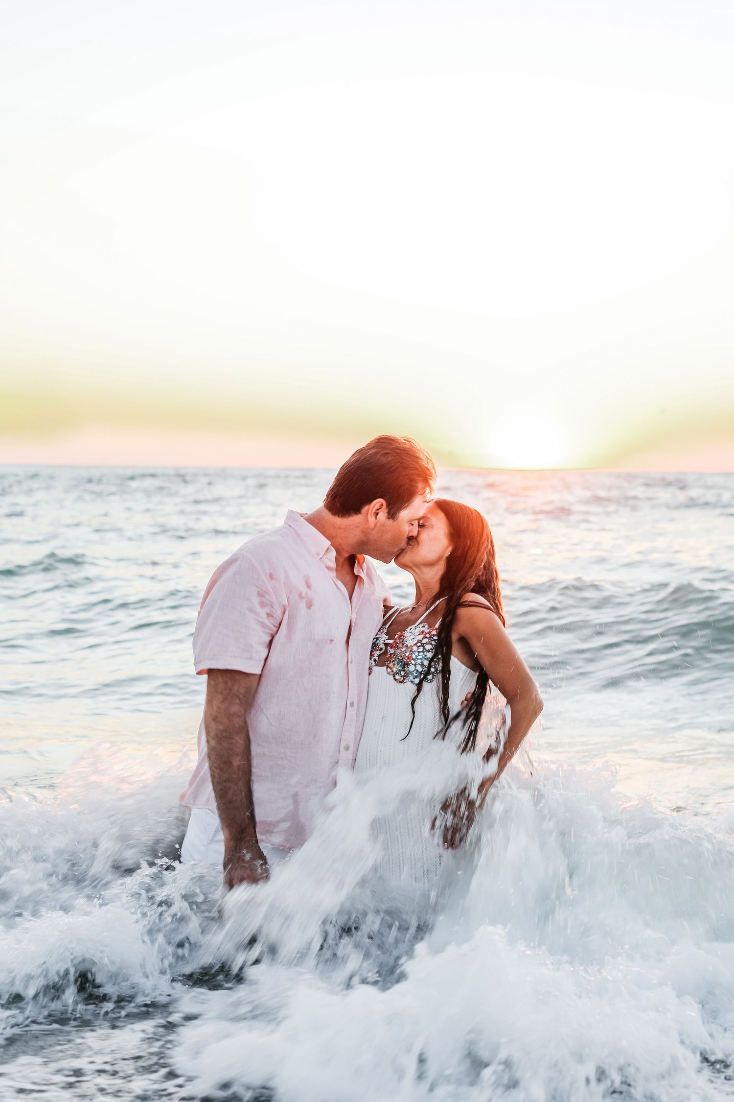 A romantic couple embraces in crashing ocean waves at sunset during a beach photo session.
