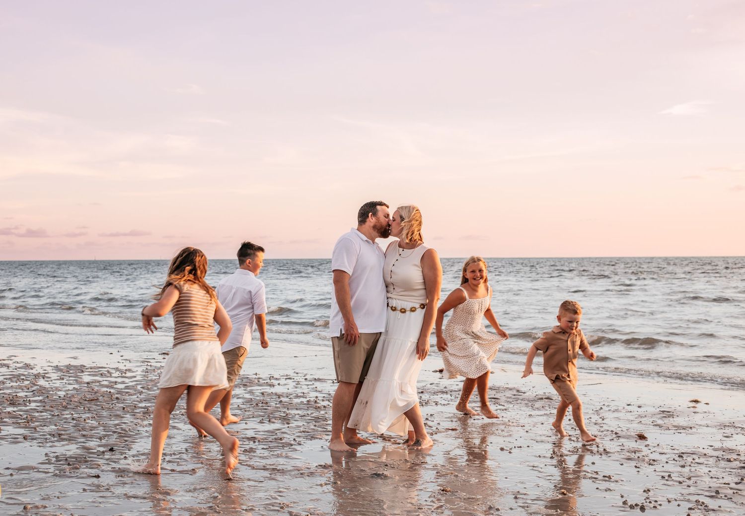 Family enjoying a golden sunset beach walk, with children playing in the waves while parents share a romantic kiss.