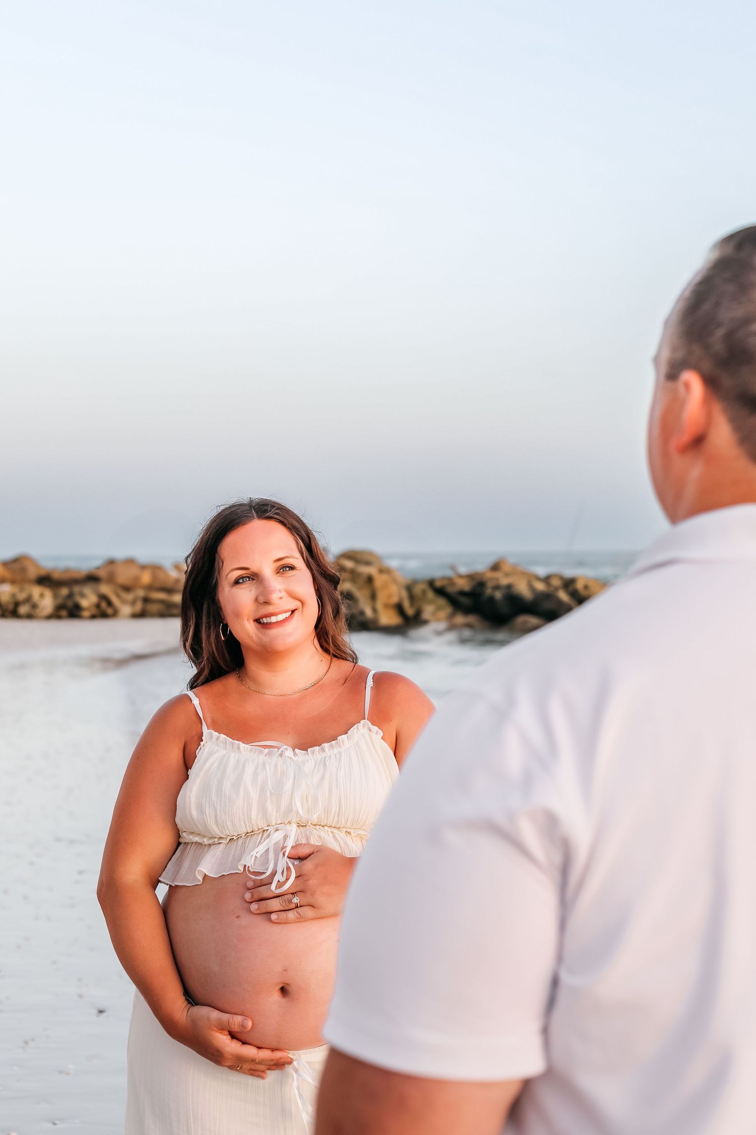 A person in a white crop top poses against a beachy coastal backdrop with rocks and ocean in the background.