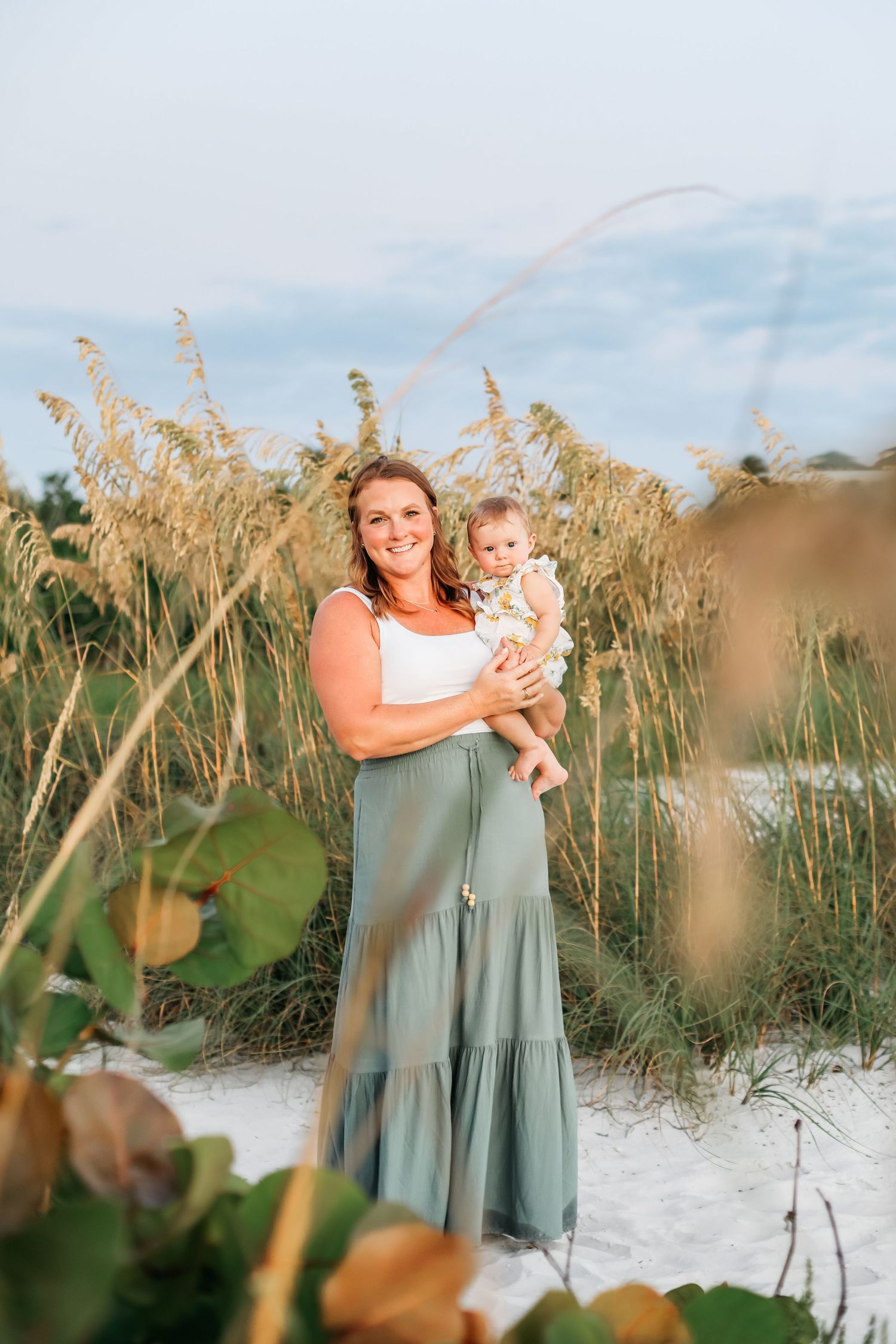 Mother and baby matching in sage green dresses against beach sea oats at golden hour.