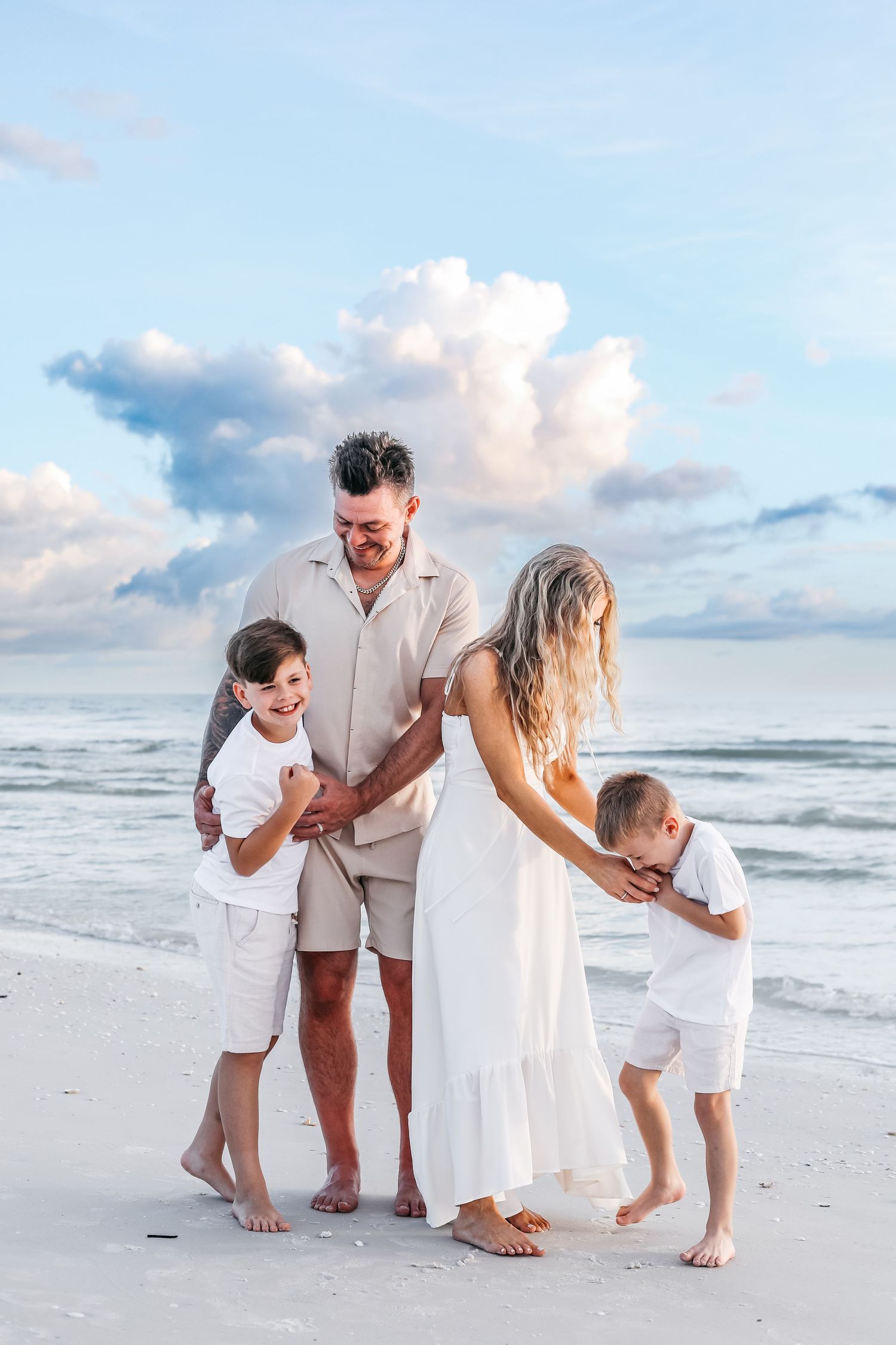 A family in white clothing poses together on a sunny beach with waves and clouds in the background.