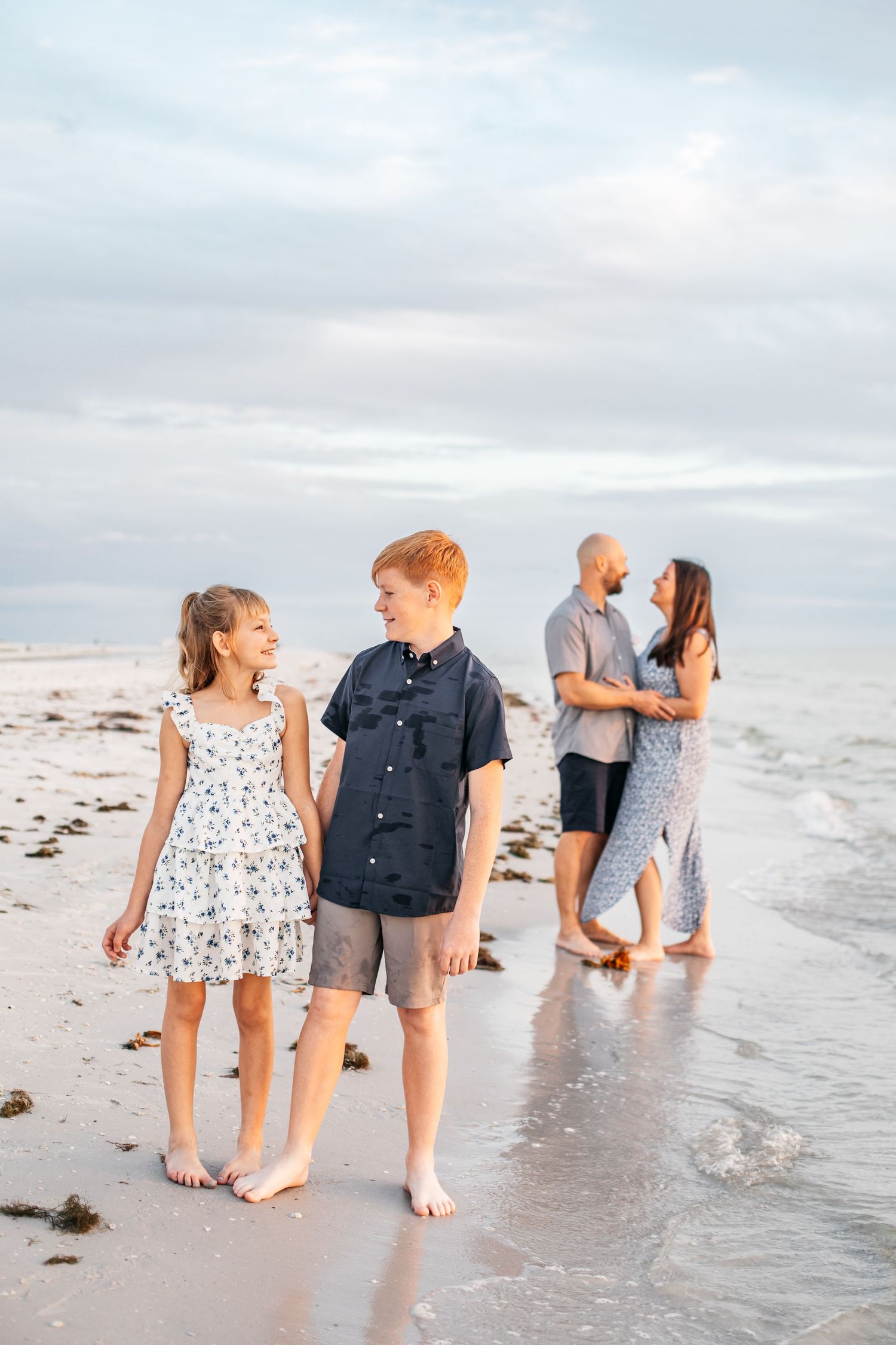 Family walking barefoot along the beach shoreline during golden hour with cloudy skies overhead.