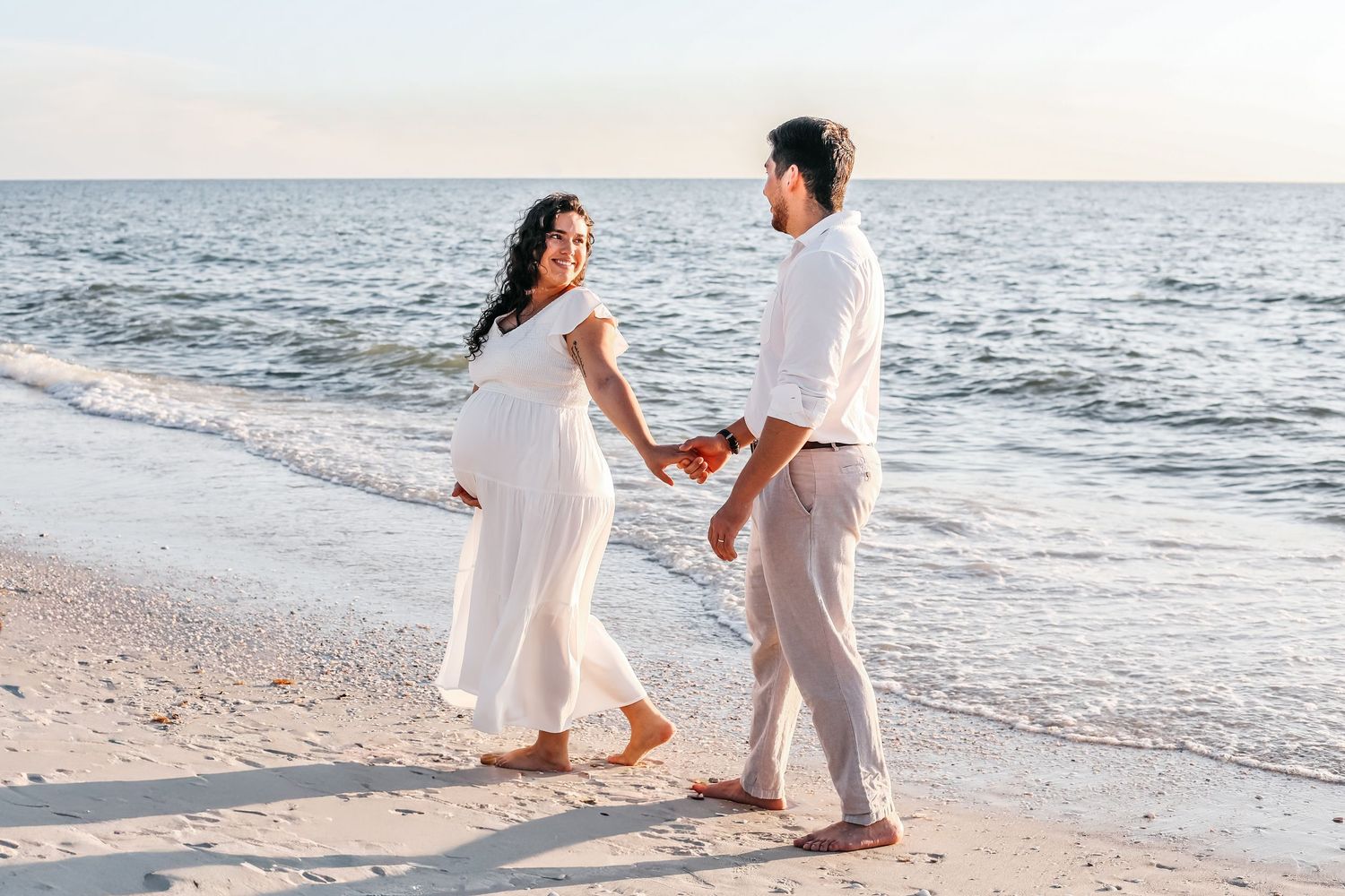 A couple in white outfits shares a romantic moment during their beach engagement photoshoot at sunset.