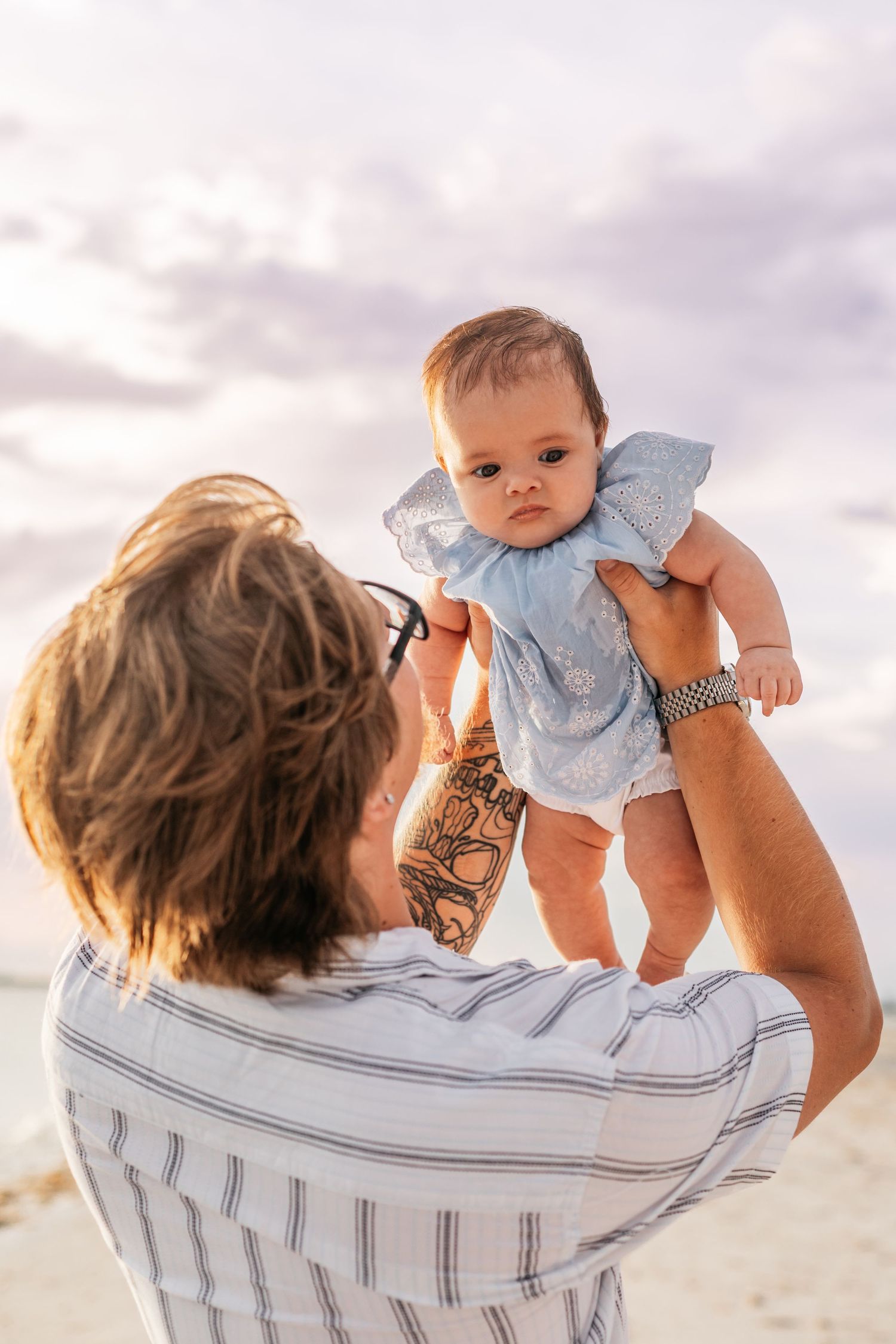 A tattooed father lifts his baby girl in a blue dress above his head on a beach at sunset.