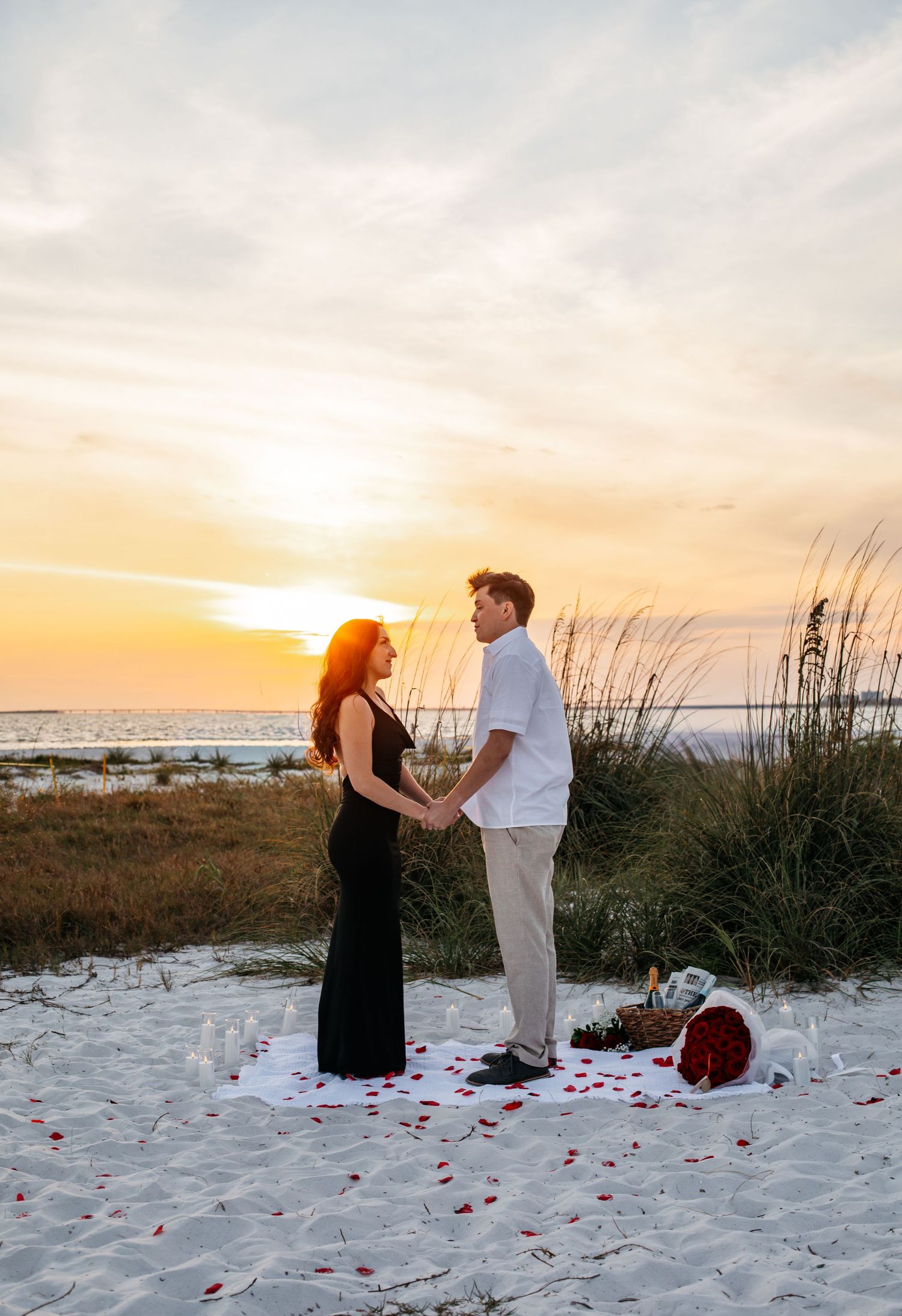 Couple holding hands on a sandy beach at sunset with rose petals and champagne glasses arranged nearby.